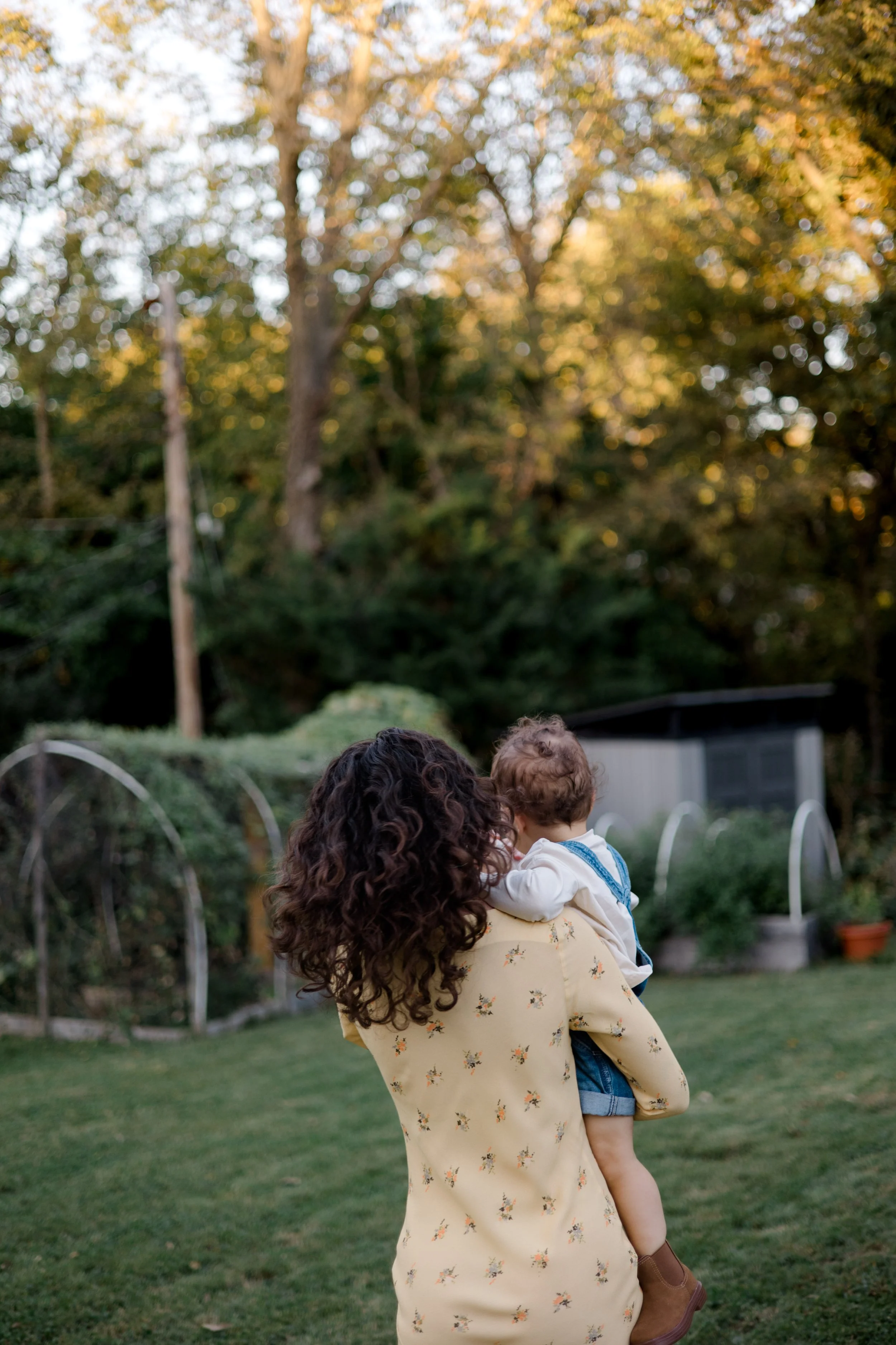 A woman with curly hair holding a small child in a backyard with trees and garden structures in the background.
