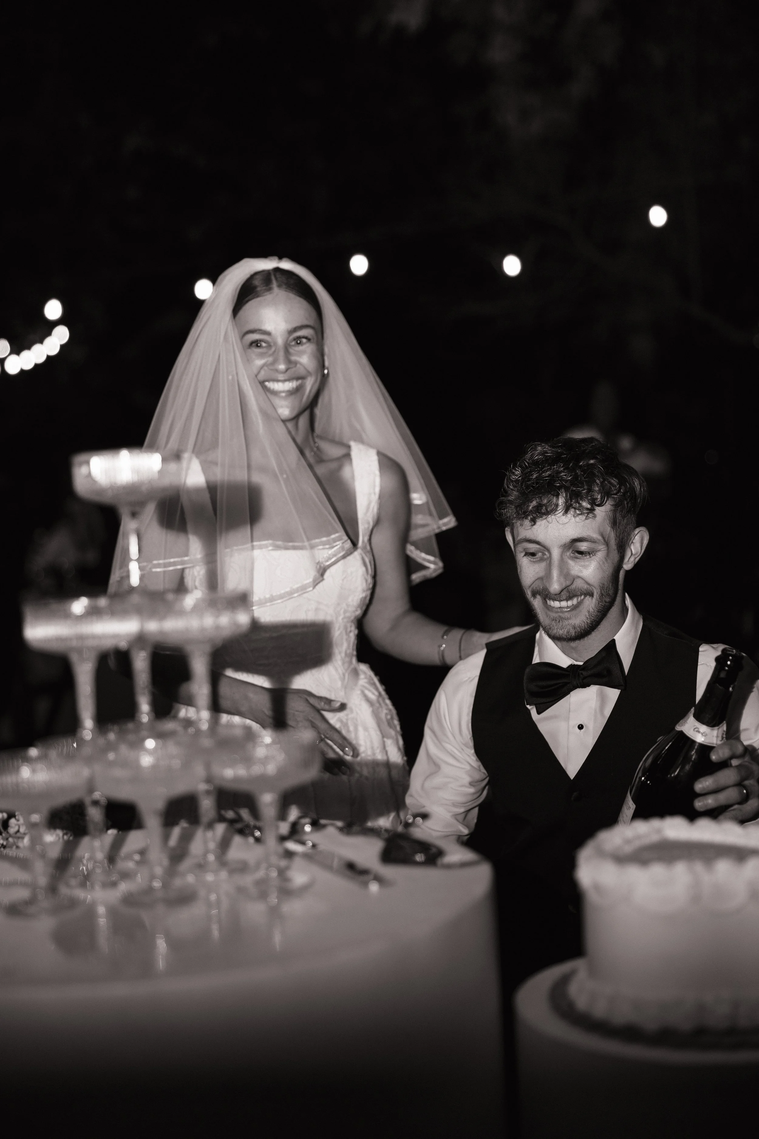 A woman in a wedding dress and veil is smiling, standing next to a man in a tuxedo with a bow tie, at a celebration or wedding reception at night. There are string lights in the background and a table with a multi-tiered cake and a bottle of champagn