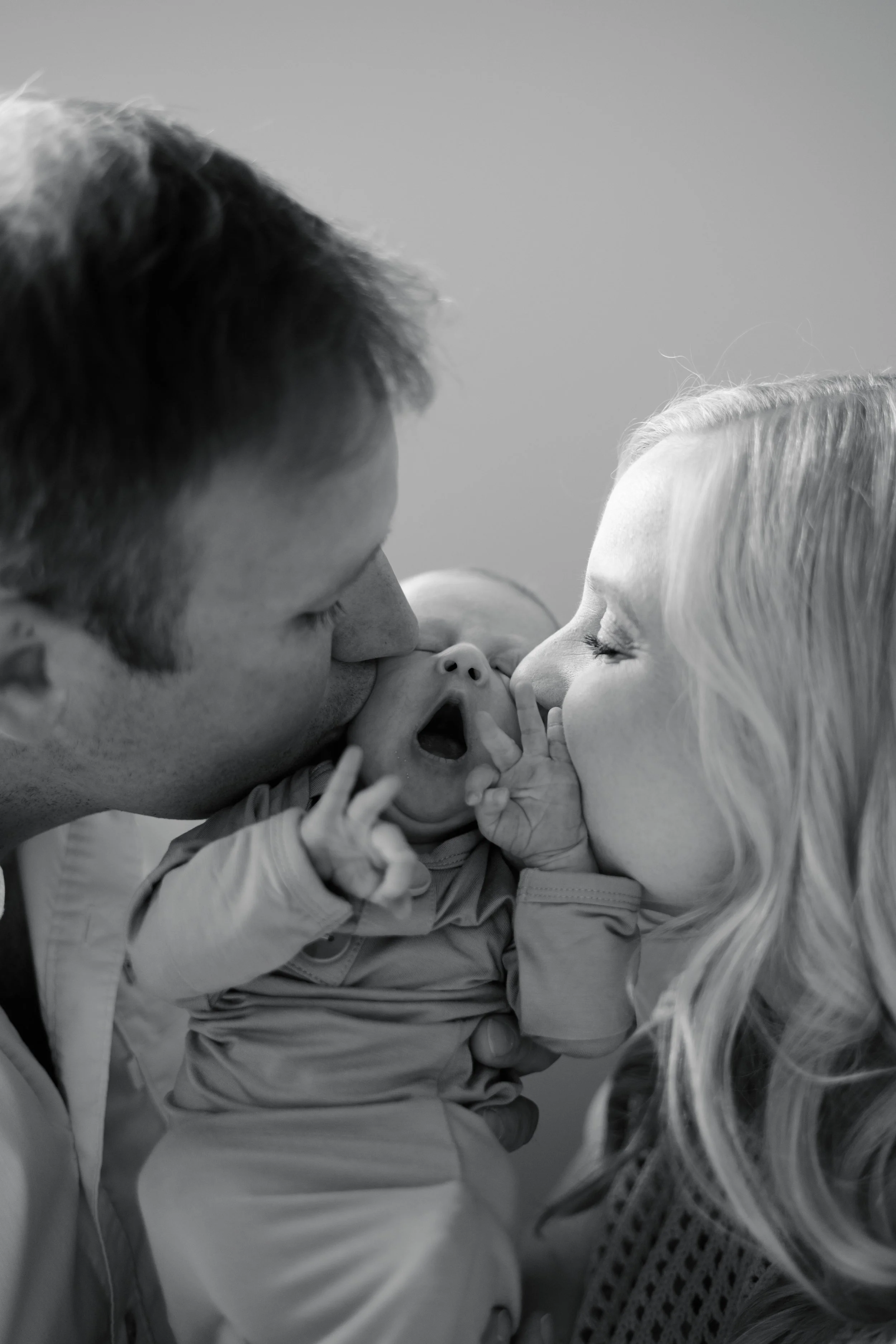 A man and a woman are kissing a baby girl, who is being cradled in their arms, in black and white.