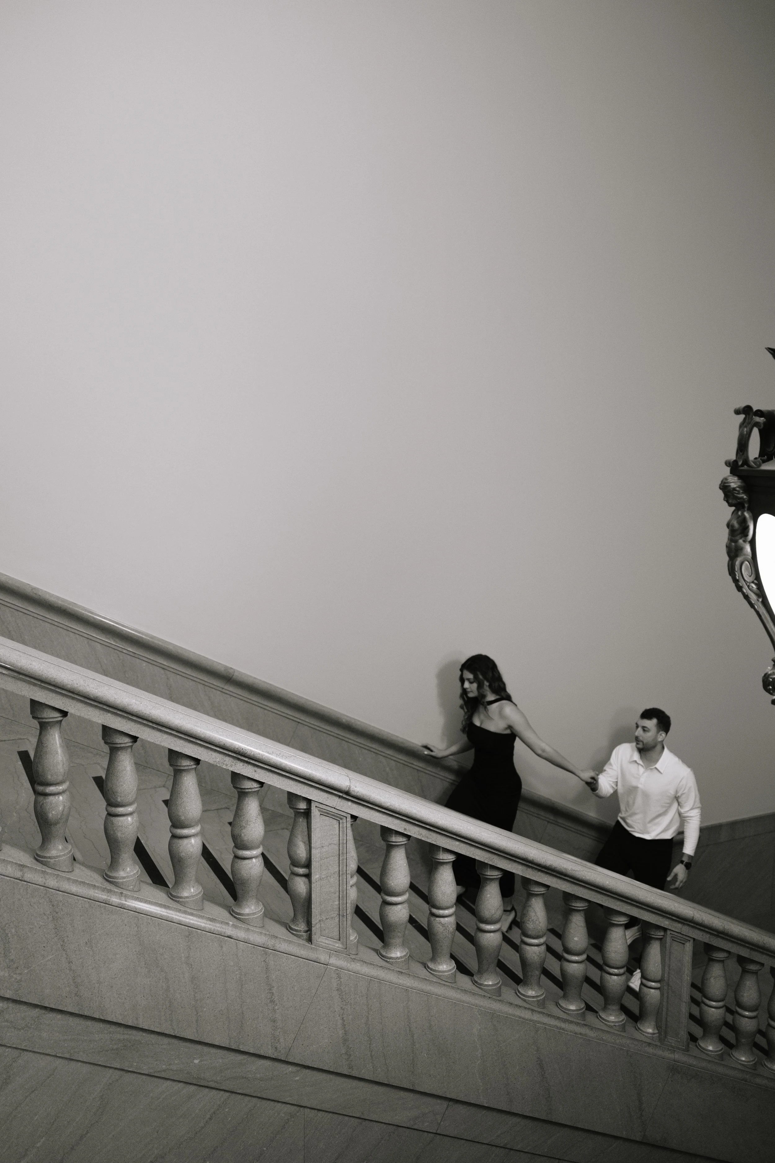 A black-and-white photo of a woman and a man holding hands and walking down a staircase indoors. The woman is wearing a black dress and the man is dressed in a light-colored shirt. The staircase has ornate balusters.