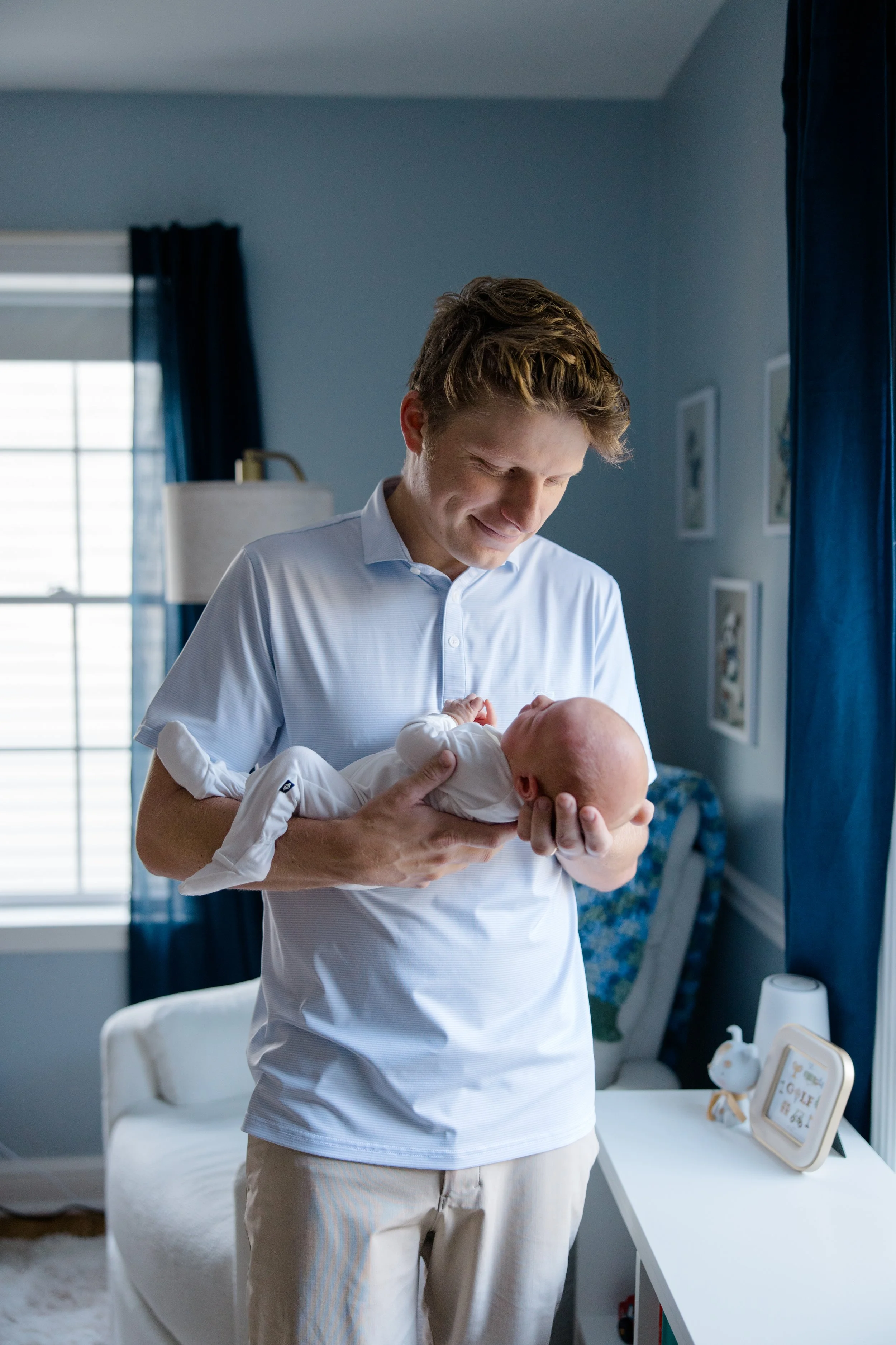A man holding a newborn baby in a blue-themed nursery, smiling and looking down at the baby.