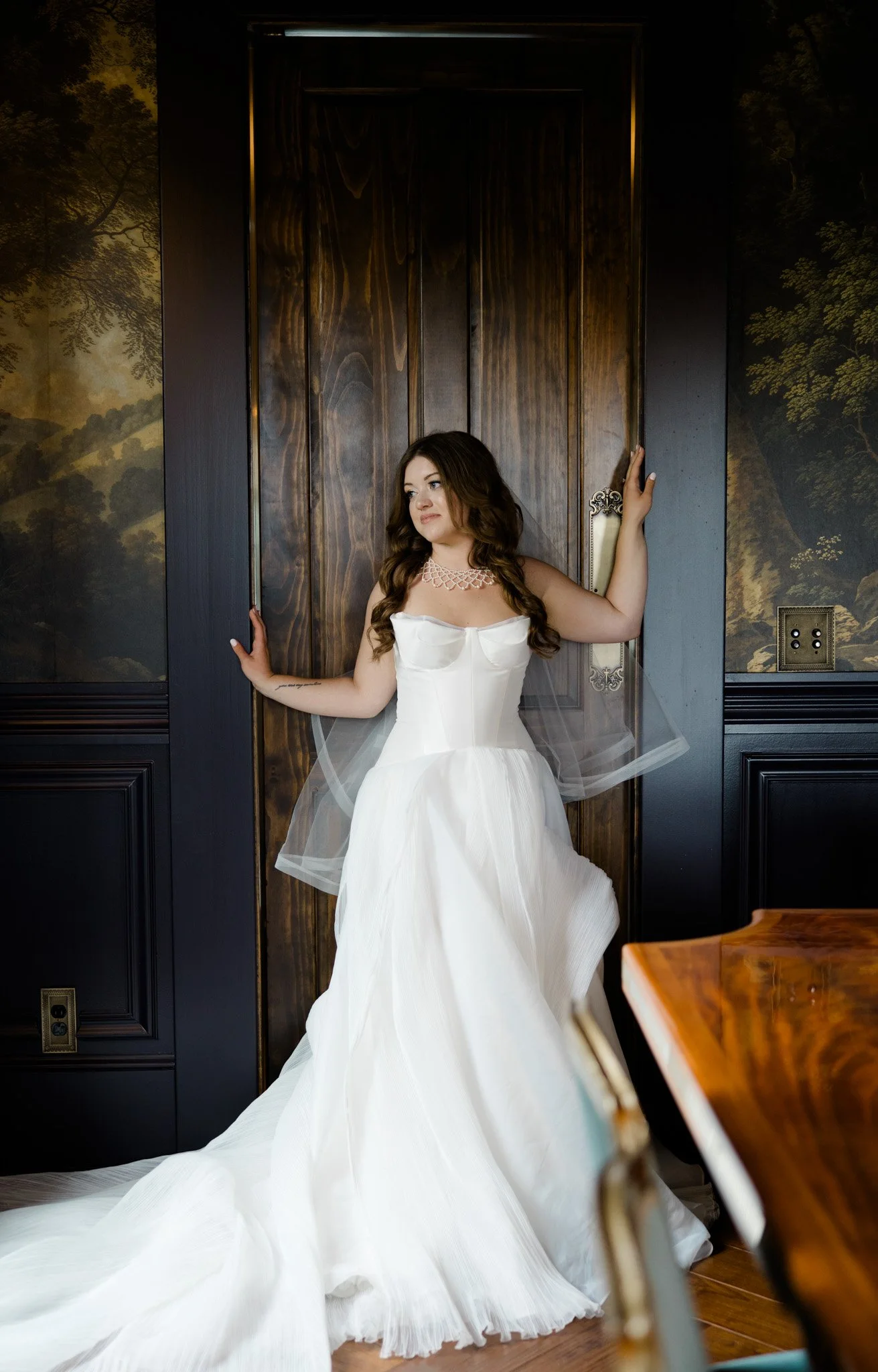A woman in a white wedding dress standing against a dark wood door with her hands on the door frame.