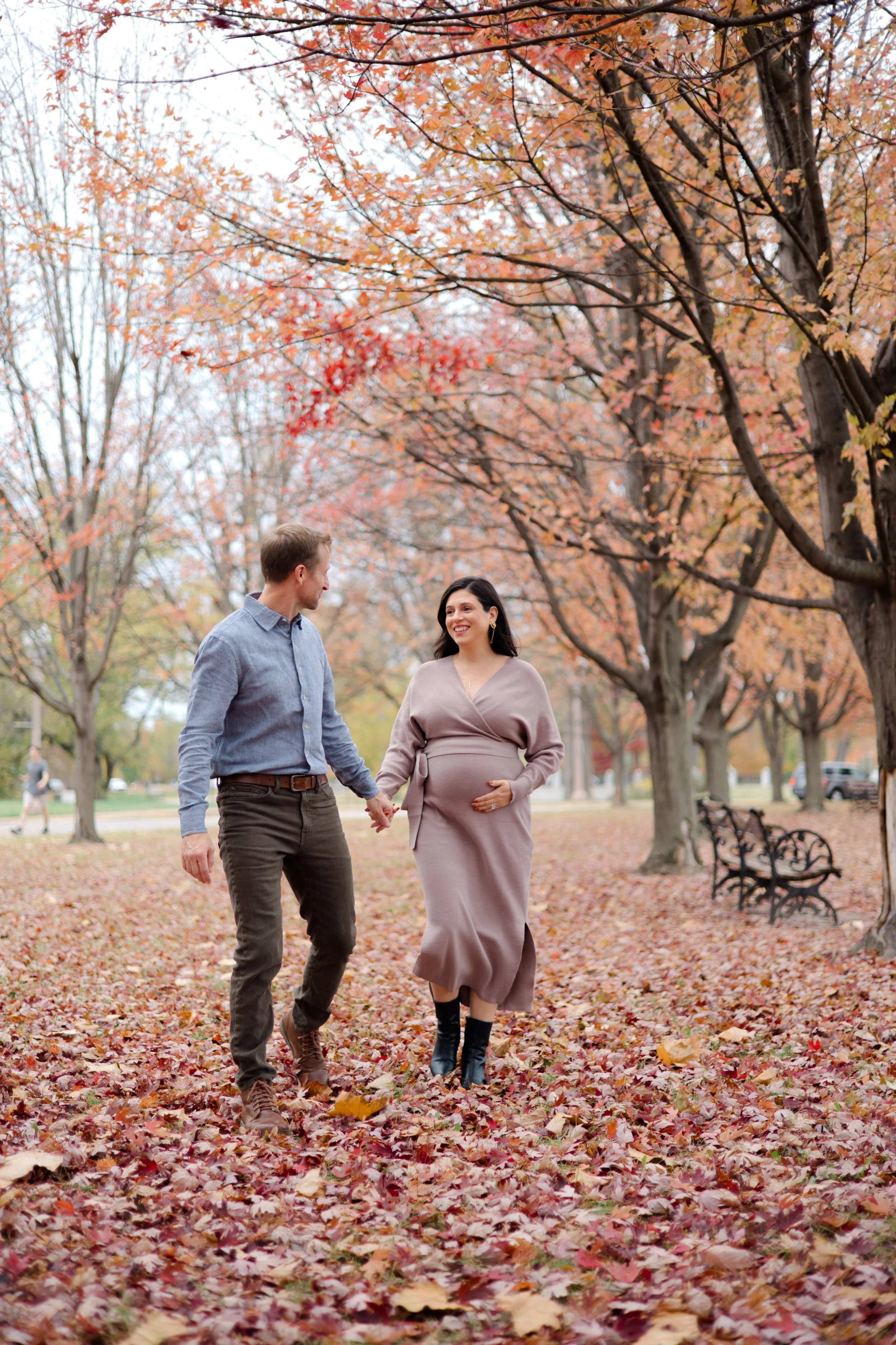 A pregnant woman and a man holding hands and walking through a park covered with fallen autumn leaves, surrounded by trees with orange and red foliage.