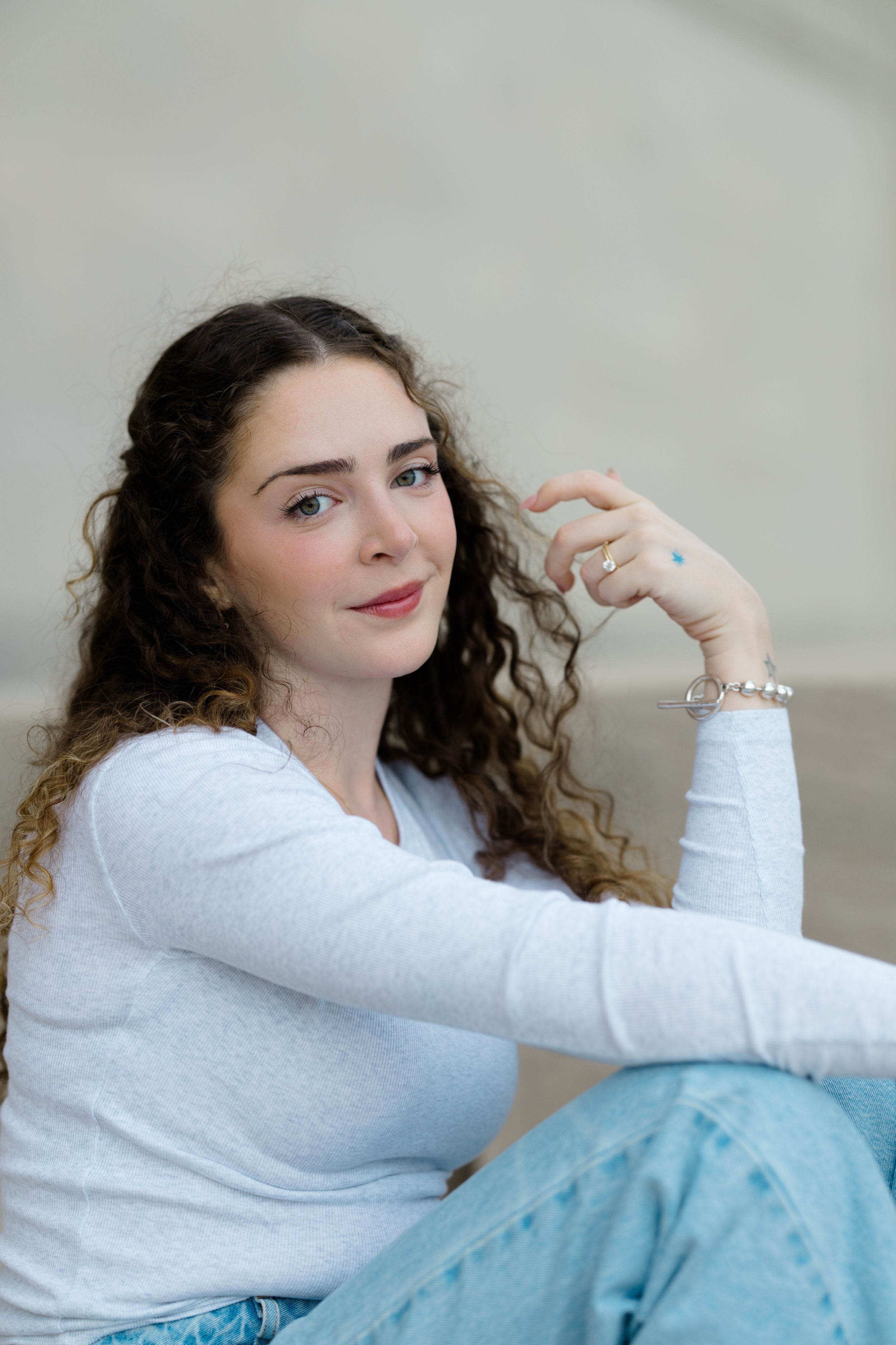 A young woman with curly brown hair and fair skin, wearing a white long-sleeve shirt and light blue jeans, sitting on the floor with her knees bent and smiling softly at the camera. She is resting her right arm on her knee and her left arm is hanging