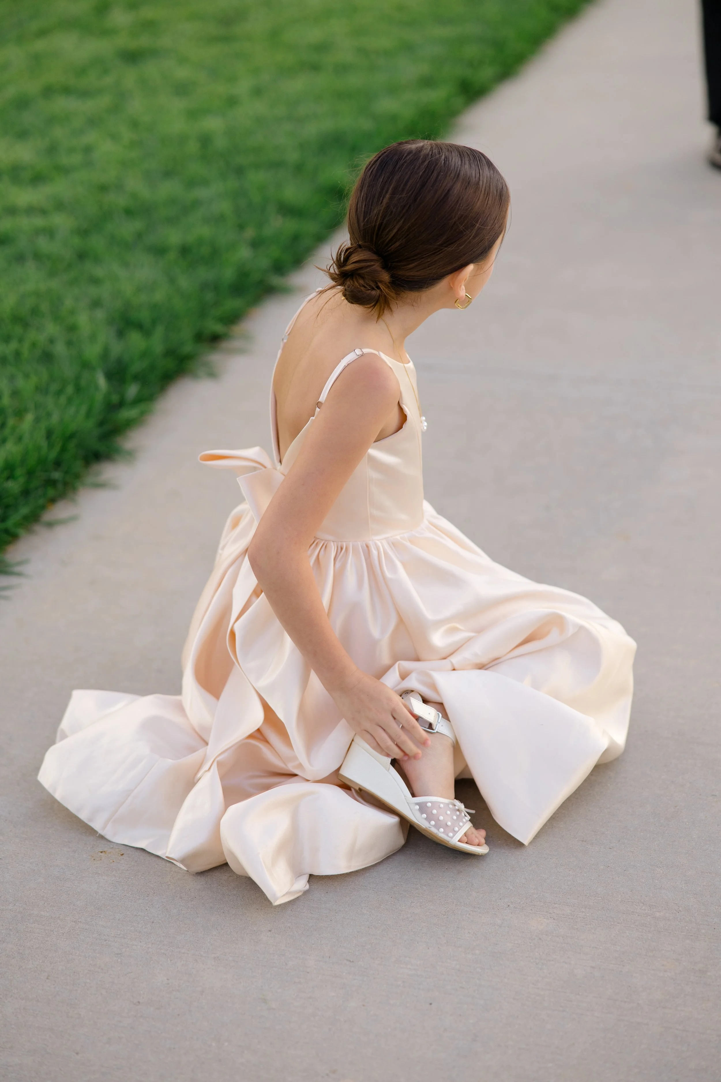 A woman dressed in a flowing, creamy-colored gown with her hair styled in a low bun, sitting down on a sidewalk near a grassy area, holding a small silver item in her hand.