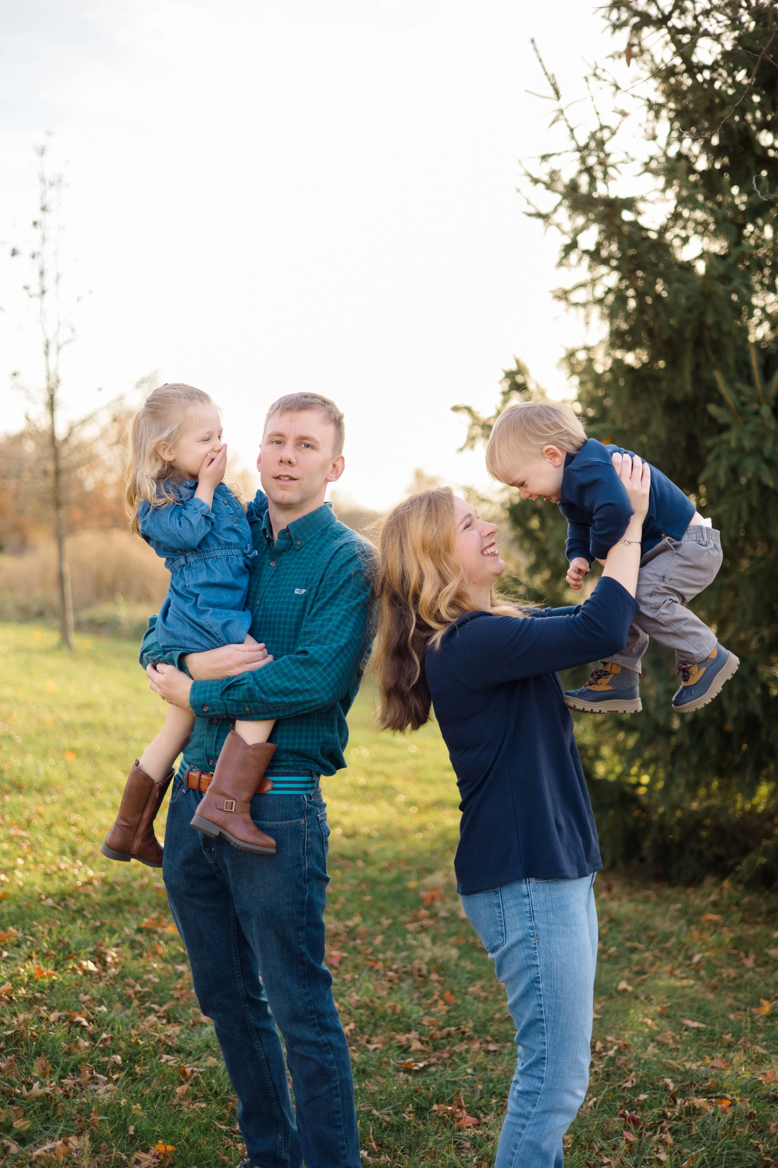 A family of four enjoying outdoor fall weather. A man is holding a young girl in his arms, and a woman is lifting a young boy in the air. All are smiling and surrounded by trees and fallen leaves.