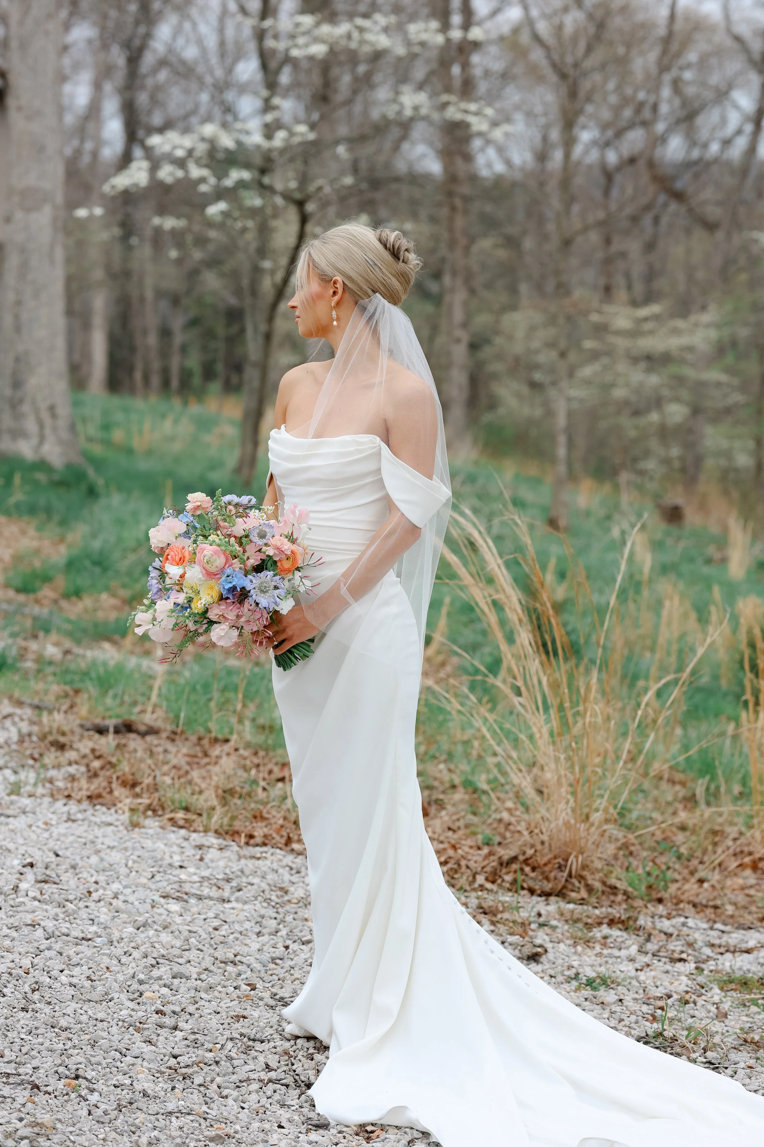 A bride in a strapless white wedding dress with a long train holding a colorful bouquet of flowers, standing outdoors in a wooded area with bare trees and green grass.