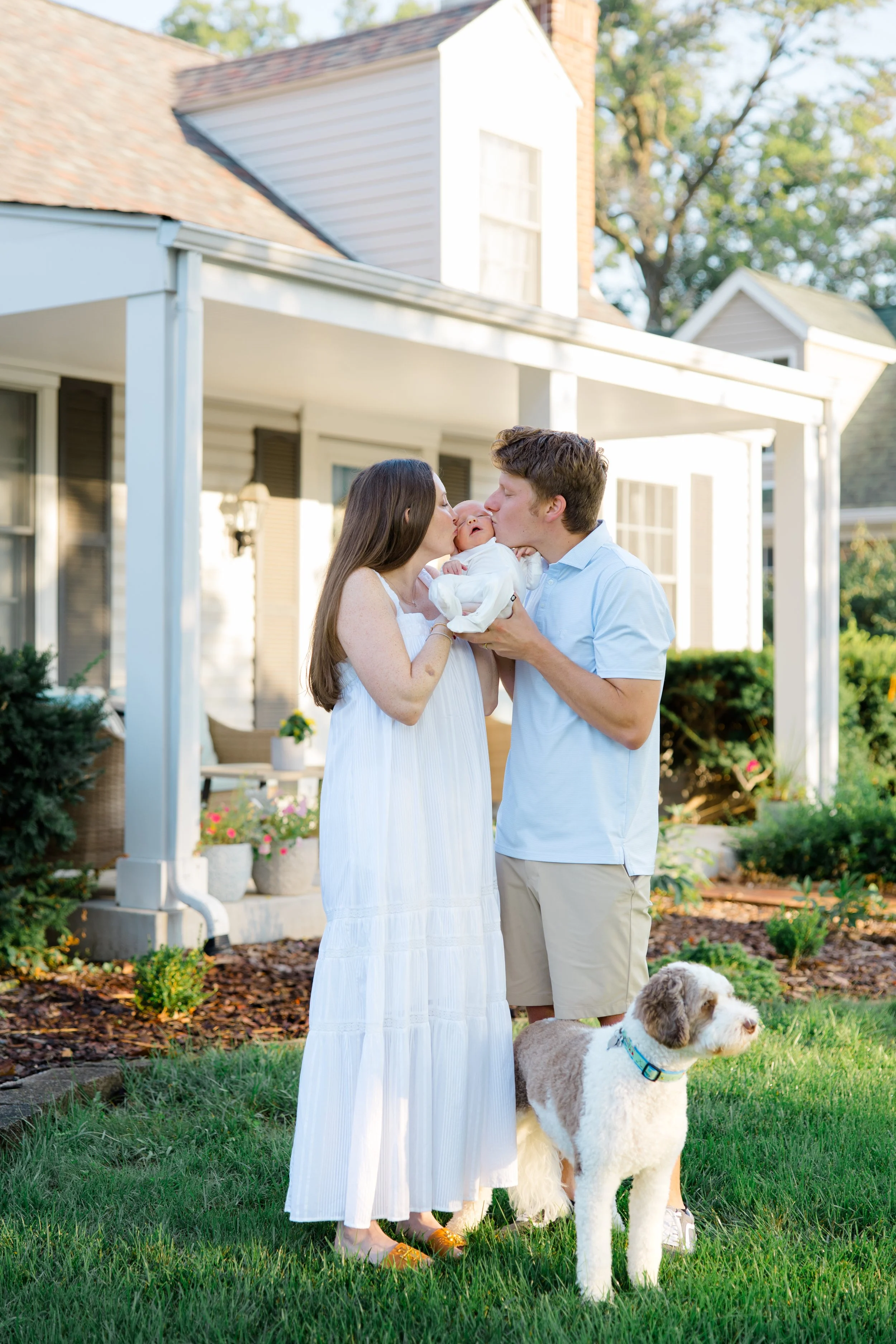 A couple holding a newborn baby and sharing a kiss outside a house with green yard and garden, with a dog nearby