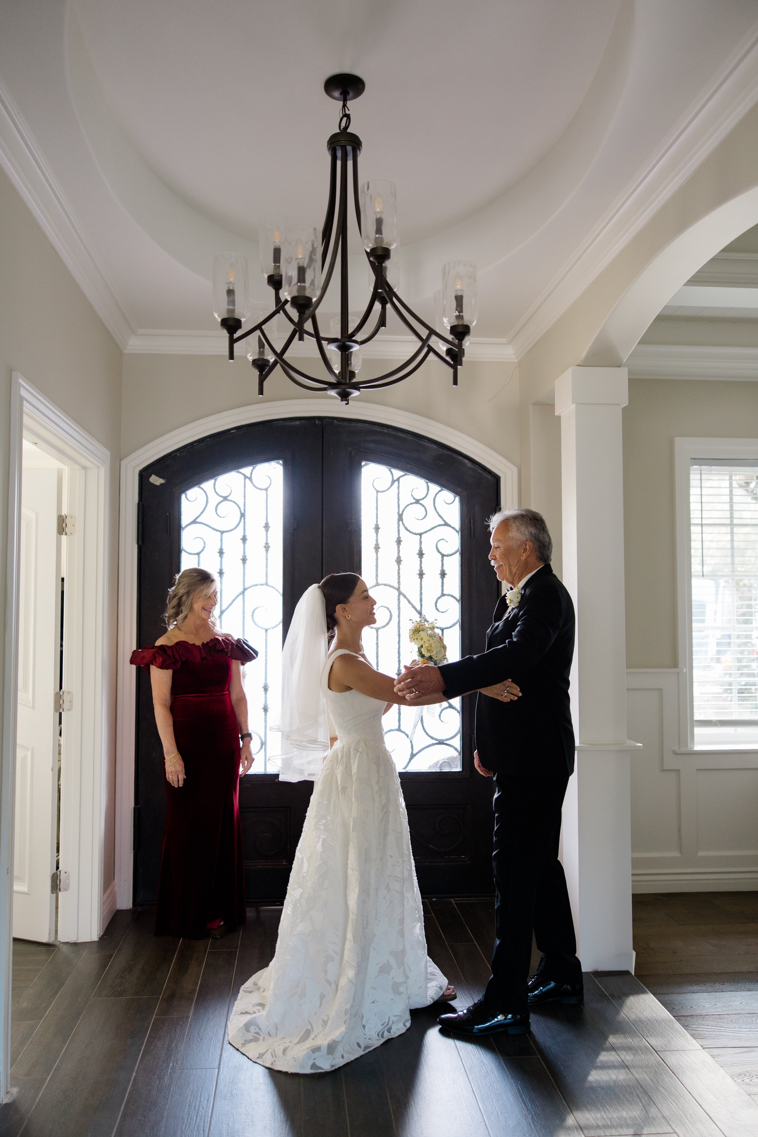 A wedding ceremony inside a house with a bride and groom holding hands and facing each other, a woman in a red dress standing behind them, and a decorative chandelier hanging from the ceiling.