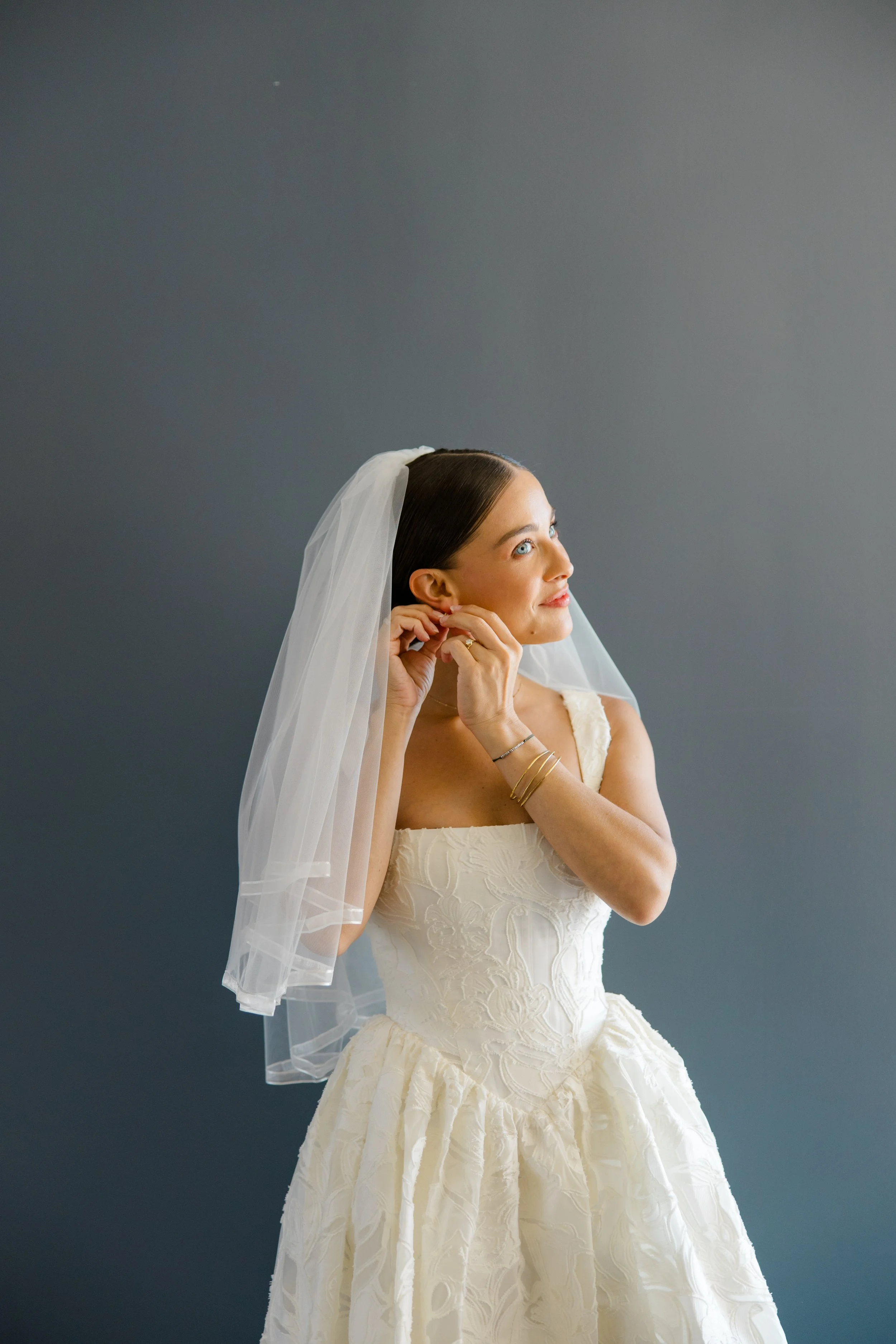 A woman in a white wedding dress adjusting her earring, with a white veil and styled hair, against a gray background.