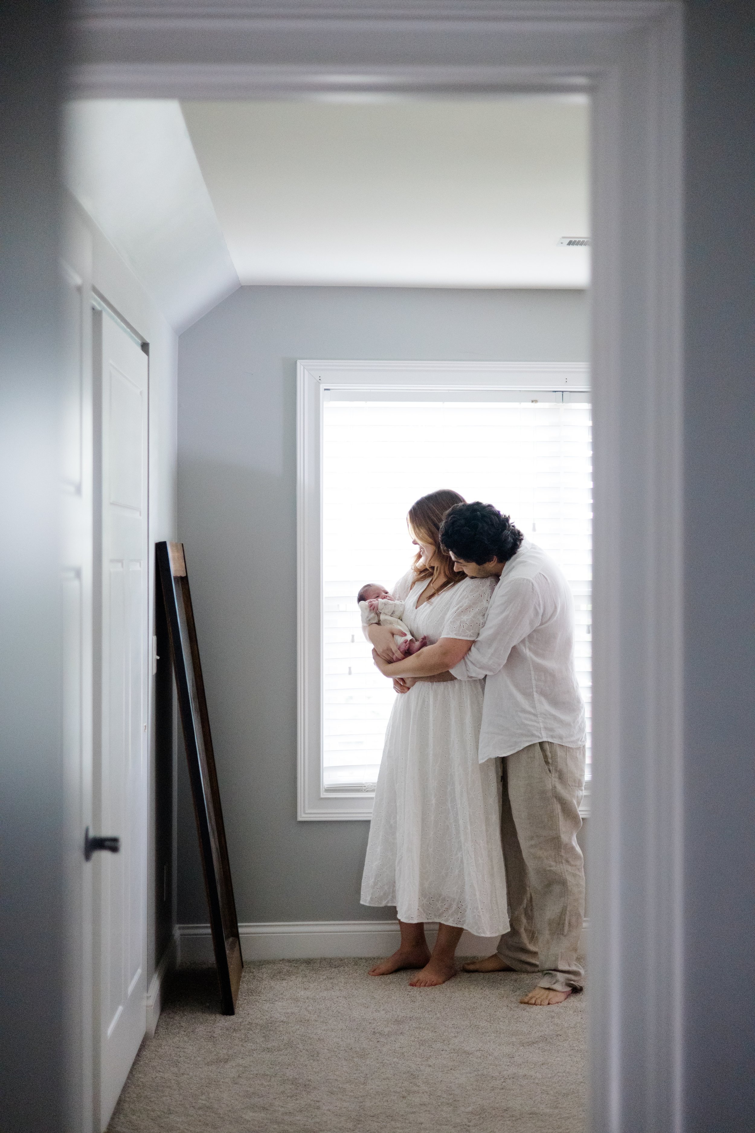 A family of three, an adult woman, adult man, and a baby, standing together in a room with a window and white blinds, as viewed through a doorway.