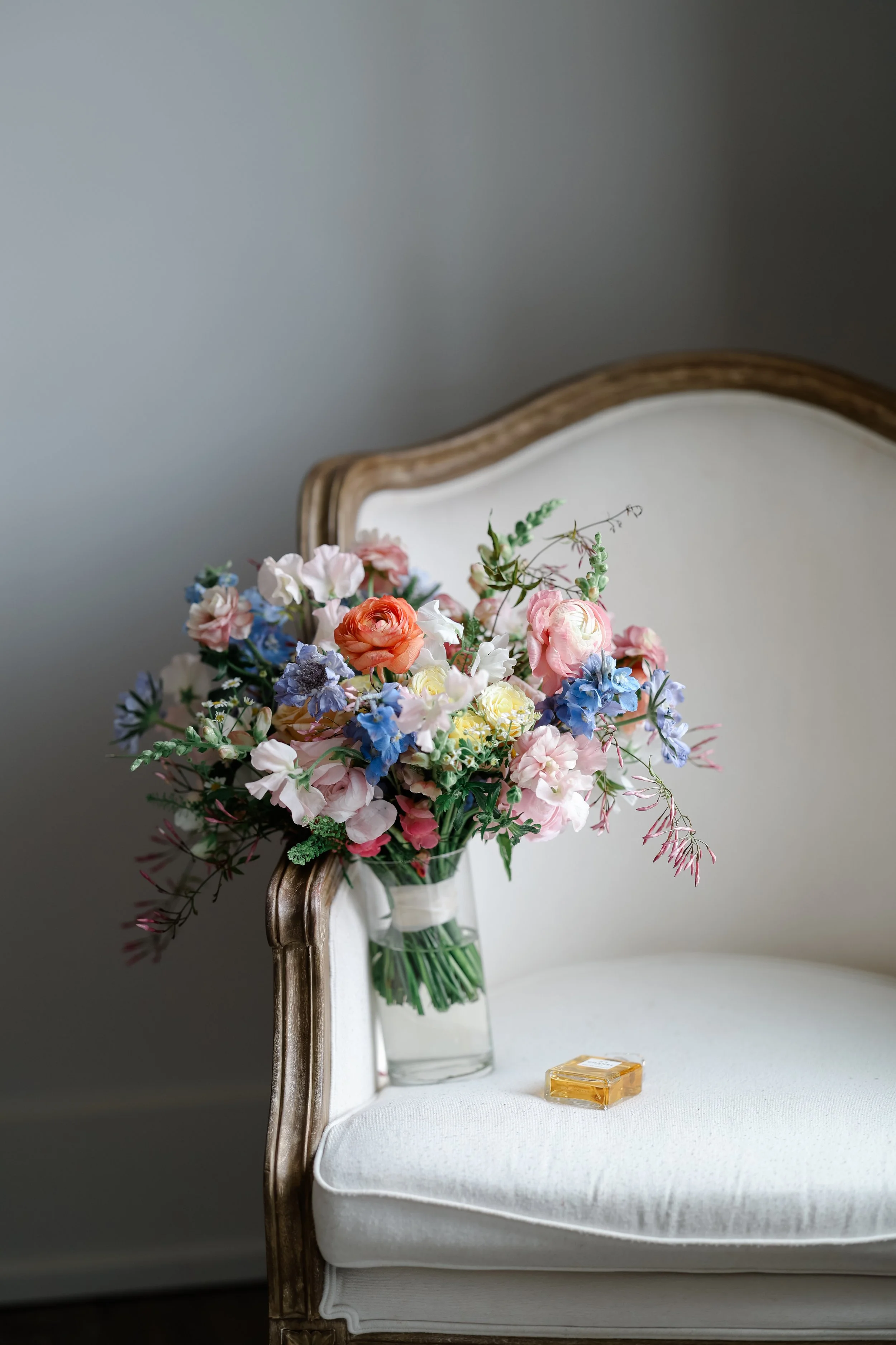 A bouquet of pink, white, peach, and blue flowers in a glass vase on a vintage white upholstered chair with wooden trim. A rectangular perfume bottle is on the chair's cushion.