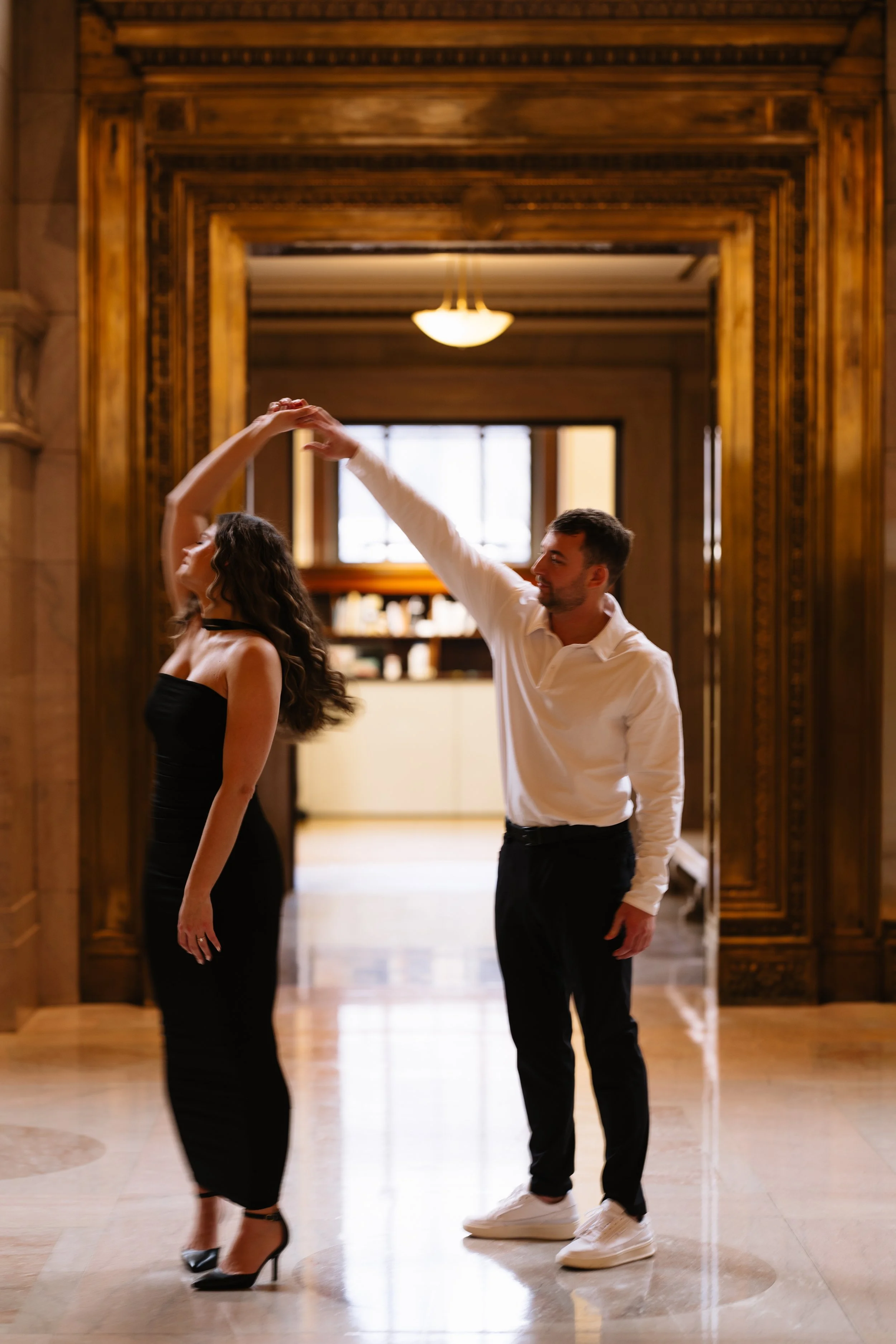 A man and woman are dancing in a grand interior with large wooden doorframes and bright windows. The woman is wearing a black dress and high heels, and the man is in a white shirt, black pants, and white sneakers.