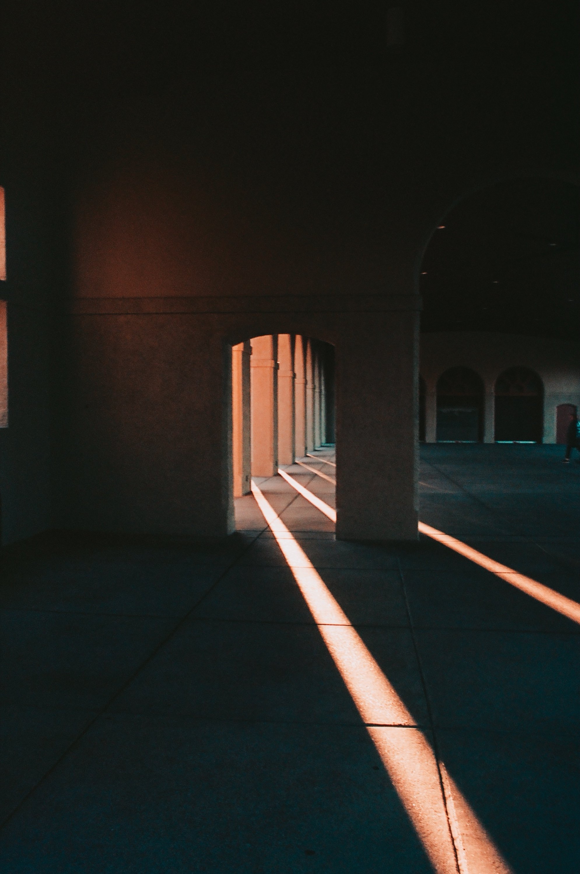 Sunlight creates a long shadow on a sidewalk in front of a building with arches, during sunset or sunrise.