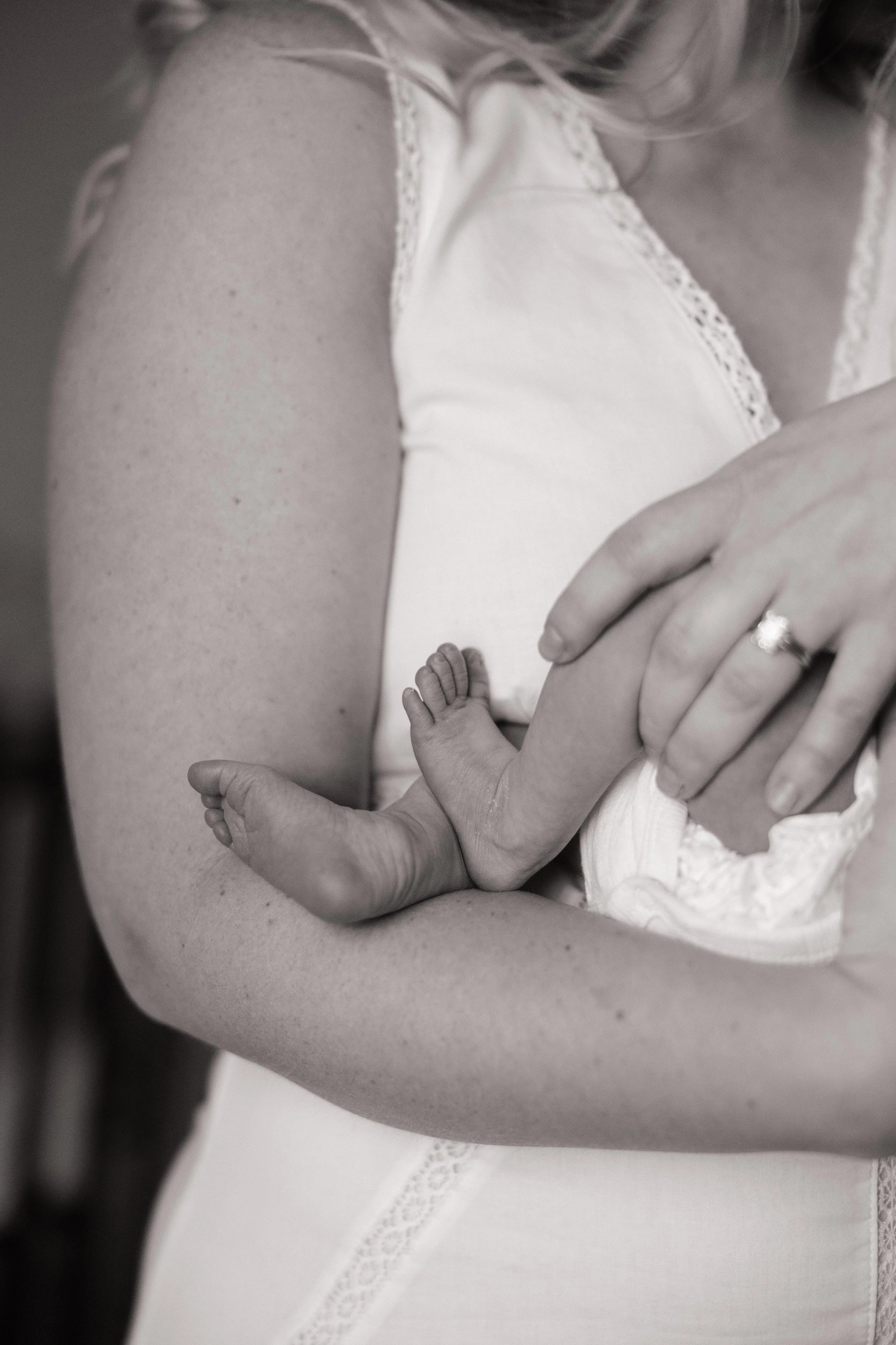 A woman holding a newborn baby close to her chest. The woman is wearing a sleeveless top with lace details, and the baby is wrapped in a swaddle.