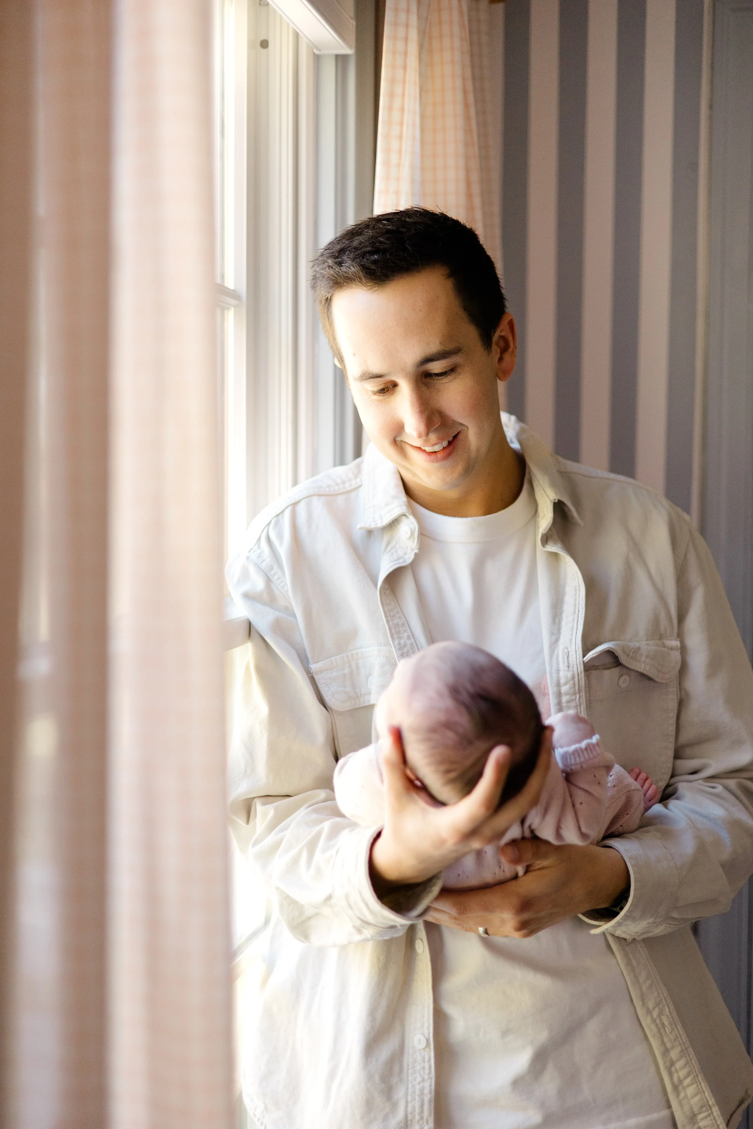 A young man with short dark hair, smiling, holding a newborn baby near a window with sunlight streaming in, in a room with striped wallpaper and curtains.