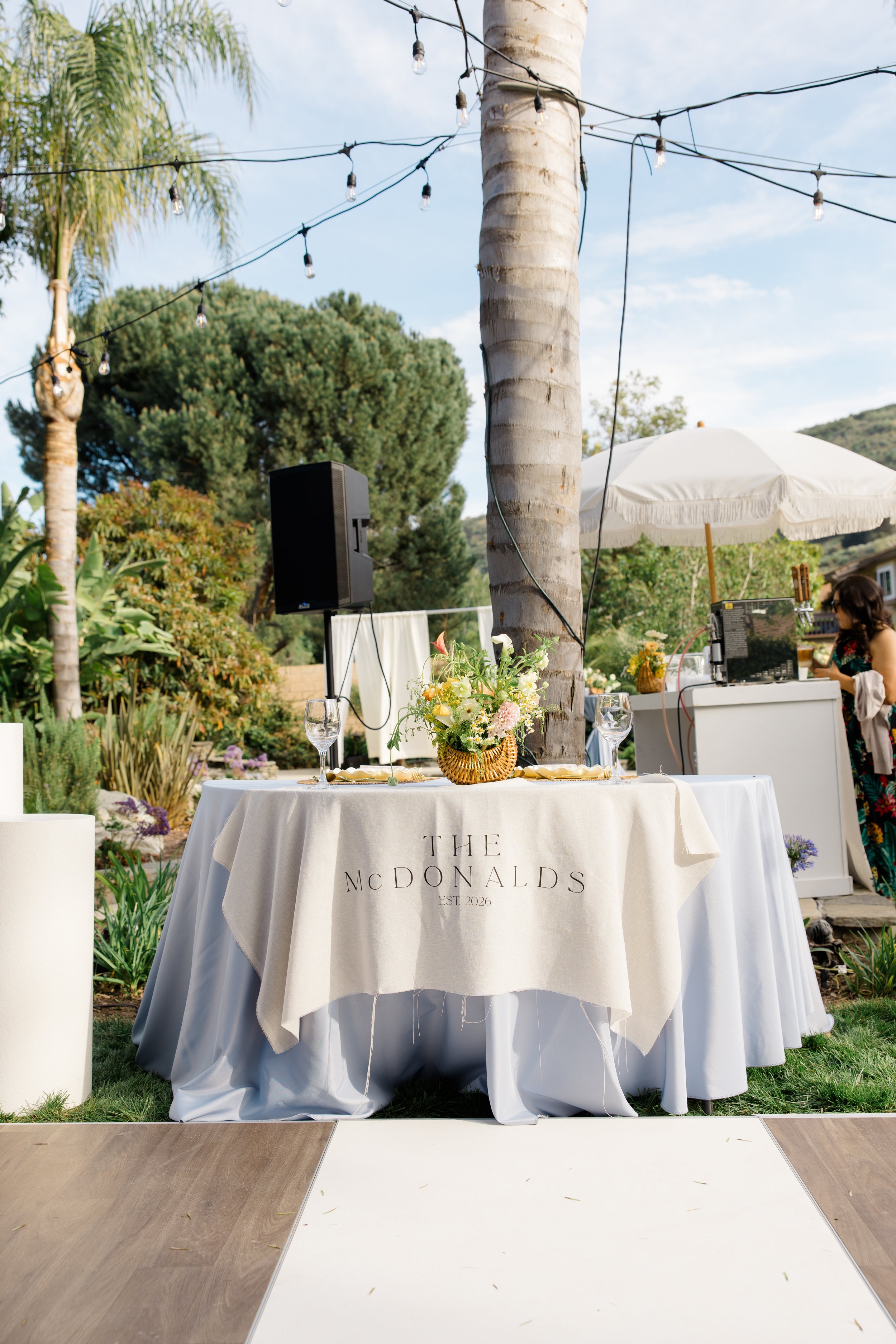 Outdoor event setup with a round table covered in a white tablecloth that has 'The McDonalds' printed on it, a floral centerpiece, empty wine glasses, and an outdoor DJ or sound setup with a laptop under a white umbrella, surrounded by trees and stri