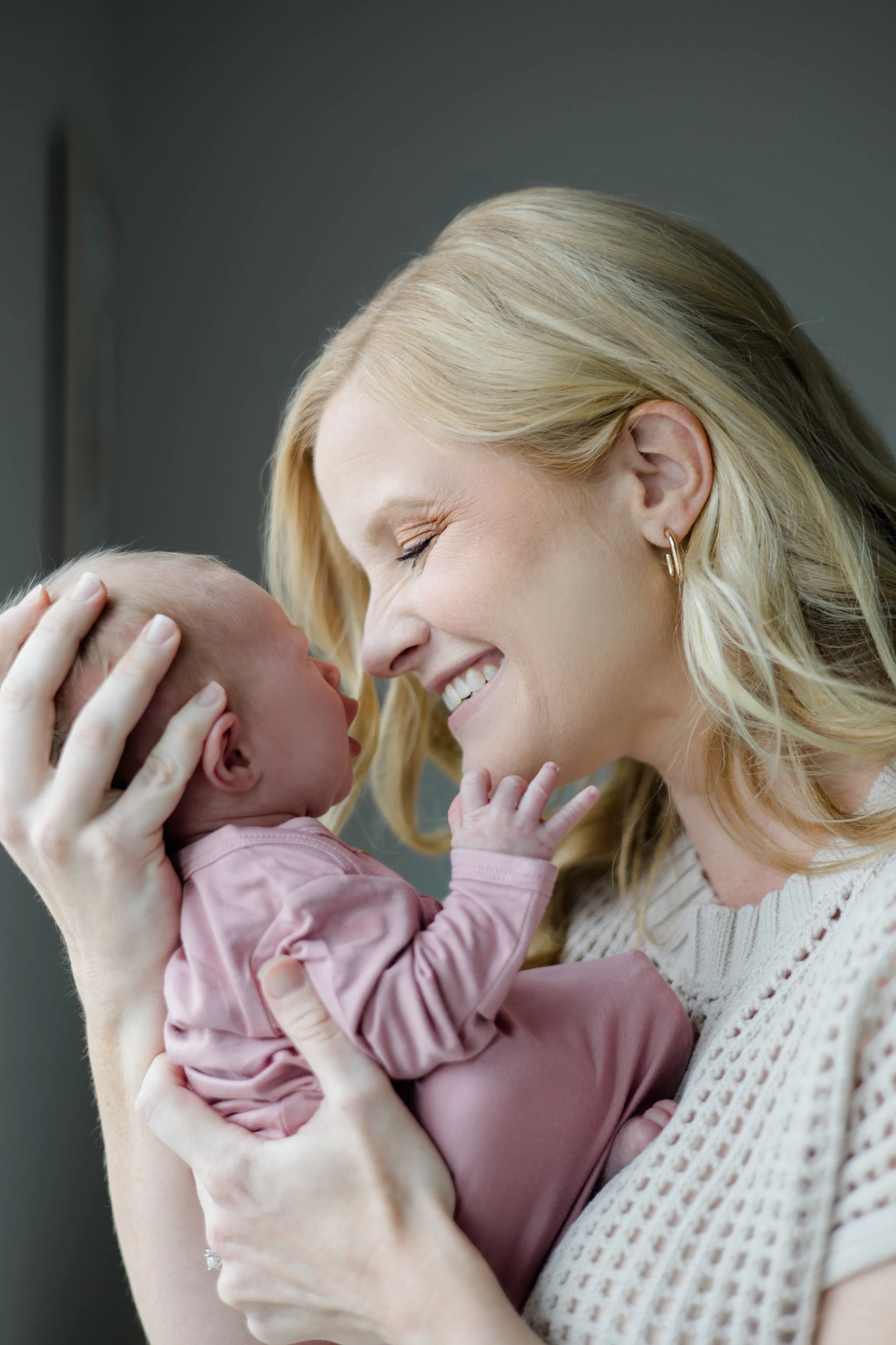 A woman with long blonde hair, wearing a cream knitted sweater, gently holds a newborn baby dressed in pink, with both smiling and touching foreheads.
