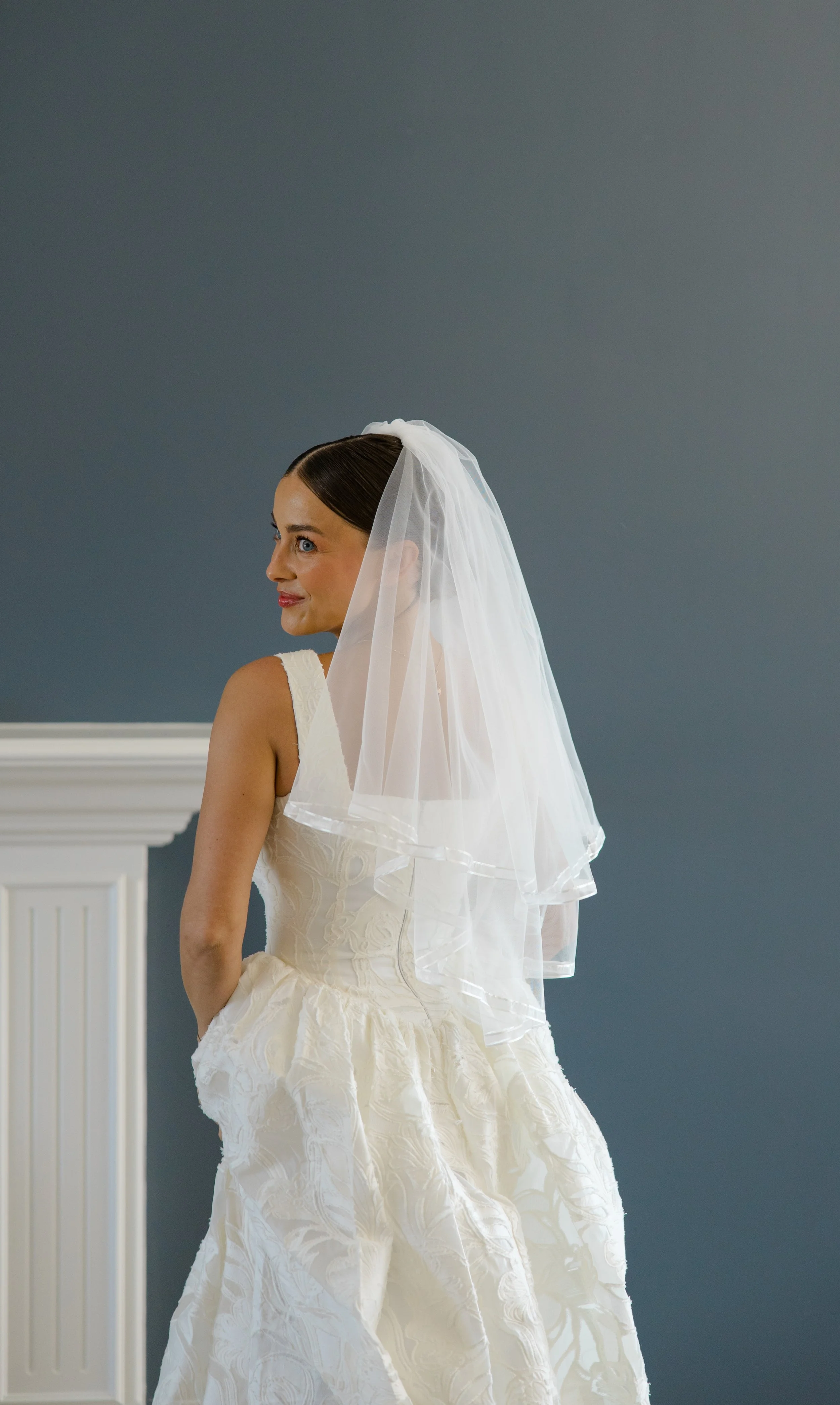 A bride with dark hair in a low bun wearing a white wedding dress with lace details and a veil, standing indoors against a blue wall with white molding.