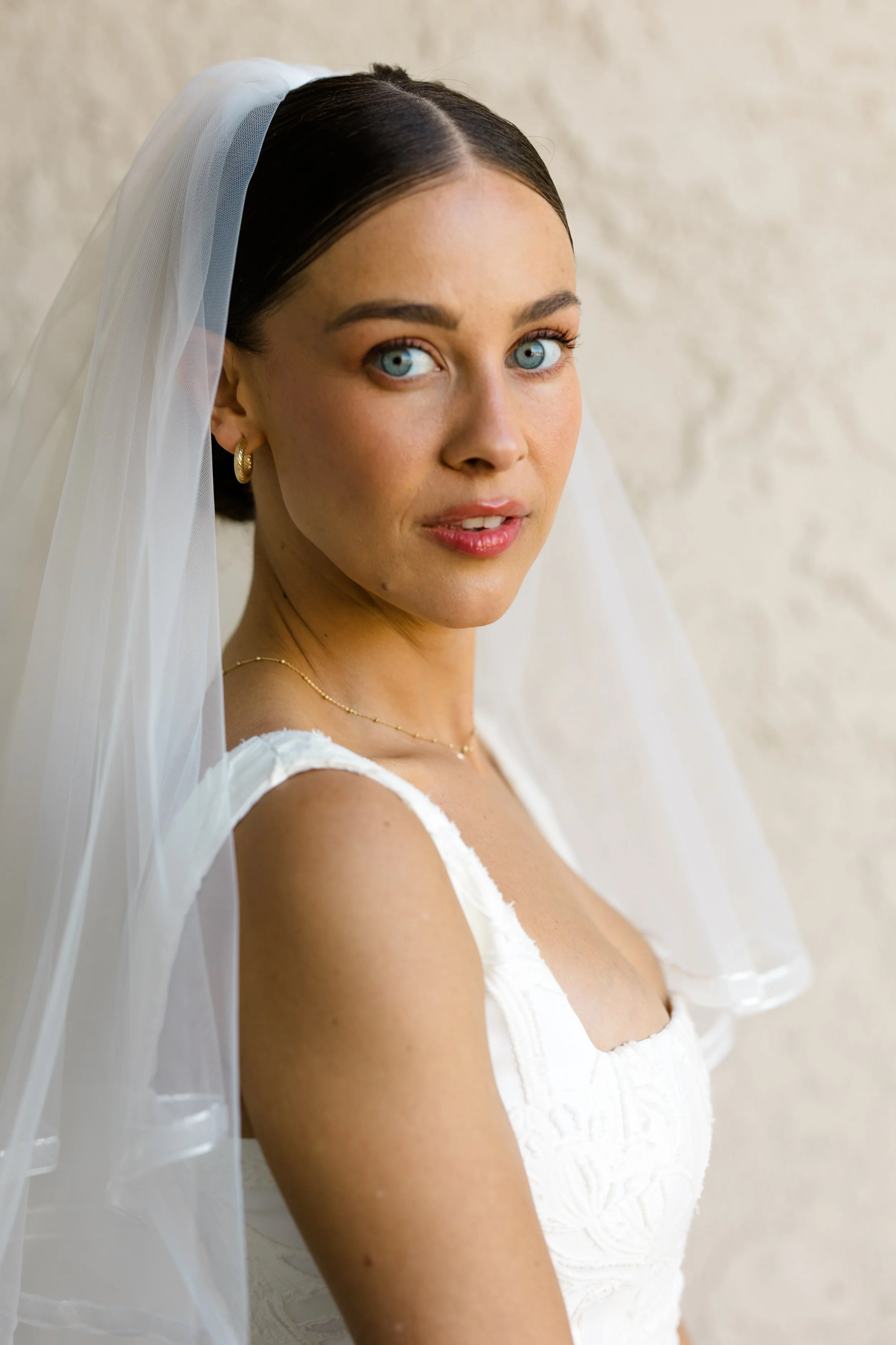 A close-up of a young woman with blue eyes, dark hair, wearing a white dress and a bridal veil, standing against a plain background.