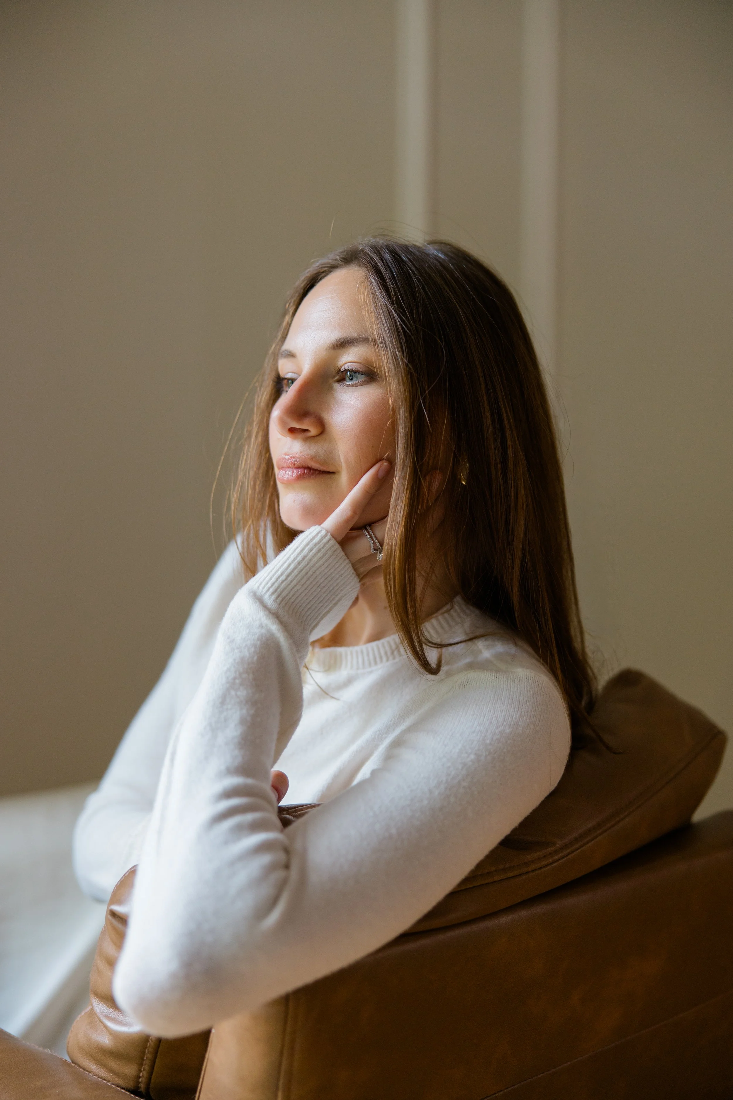 A woman with long brown hair sitting on a brown couch, wearing a white sweater, resting her chin on her hand, looking peacefully out of frame.