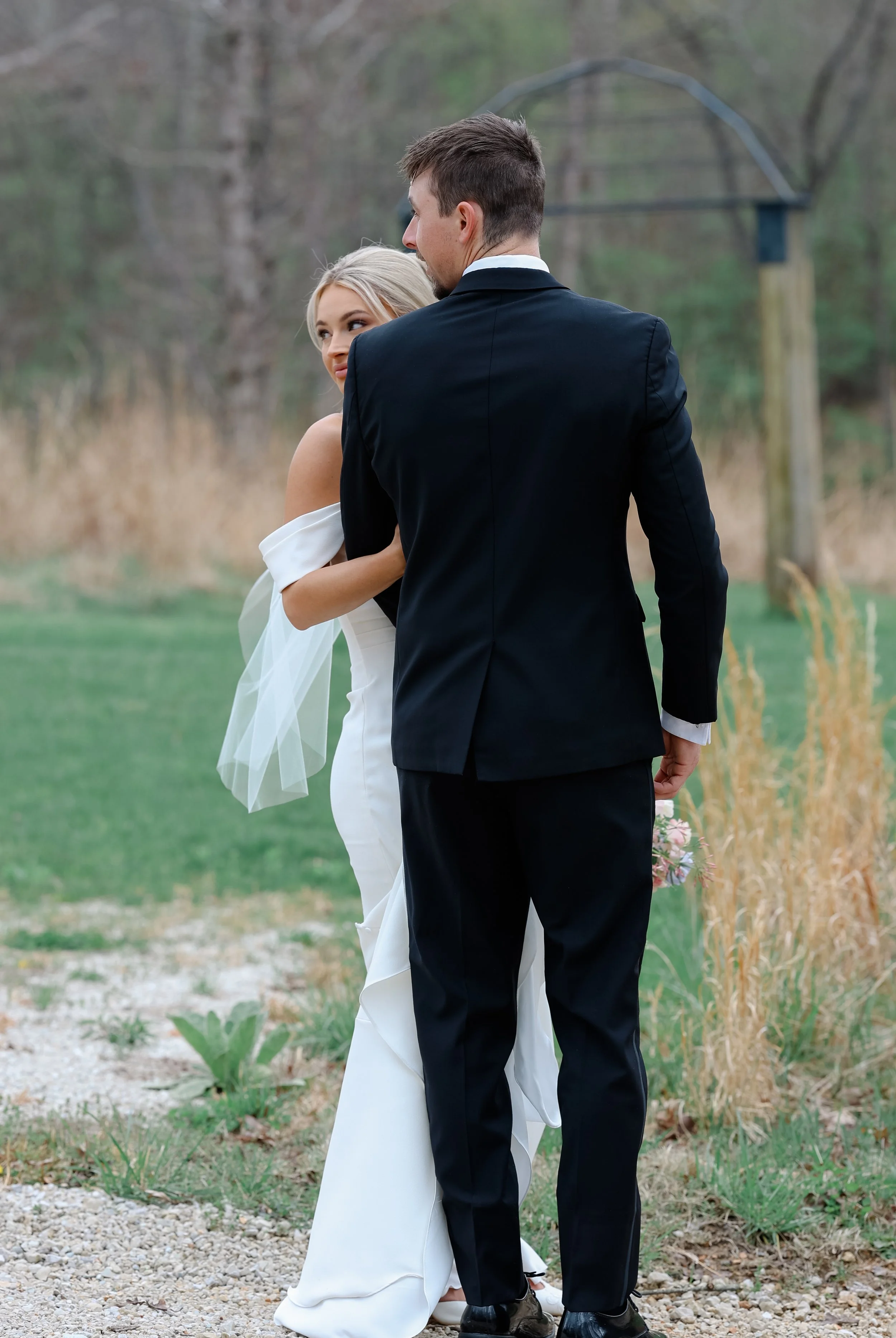 A couple in wedding attire stands outdoors on a gravel path, with the groom facing away and the bride looking at him, in a wooded area with tall grasses and trees in the background.