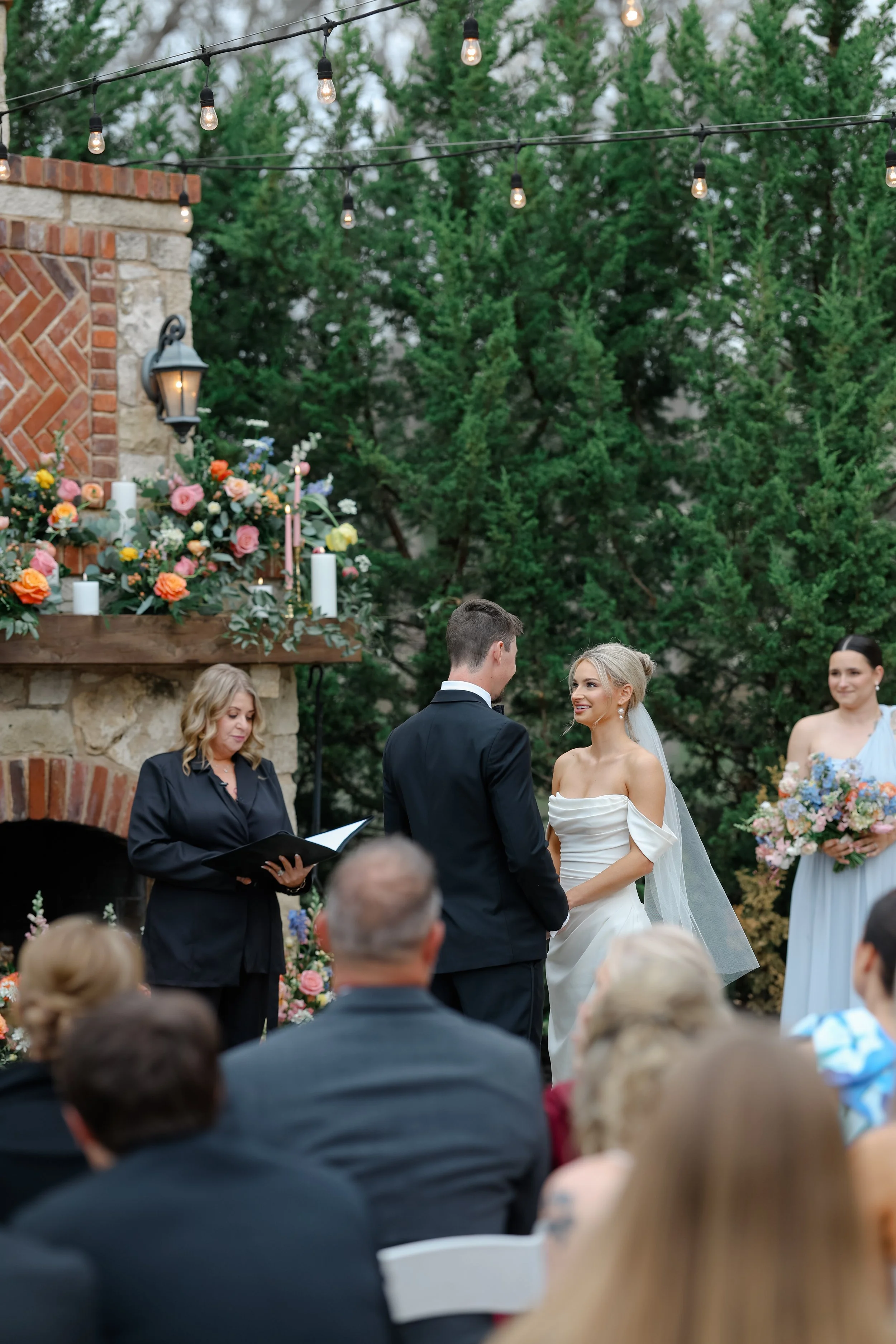 A wedding ceremony outdoors with an officiant reading from a black folder, a bride and groom facing each other, and a maid of honor holding a bouquet of flowers. Guests are seated in chairs, and there are string lights, a brick wall, and greenery in 