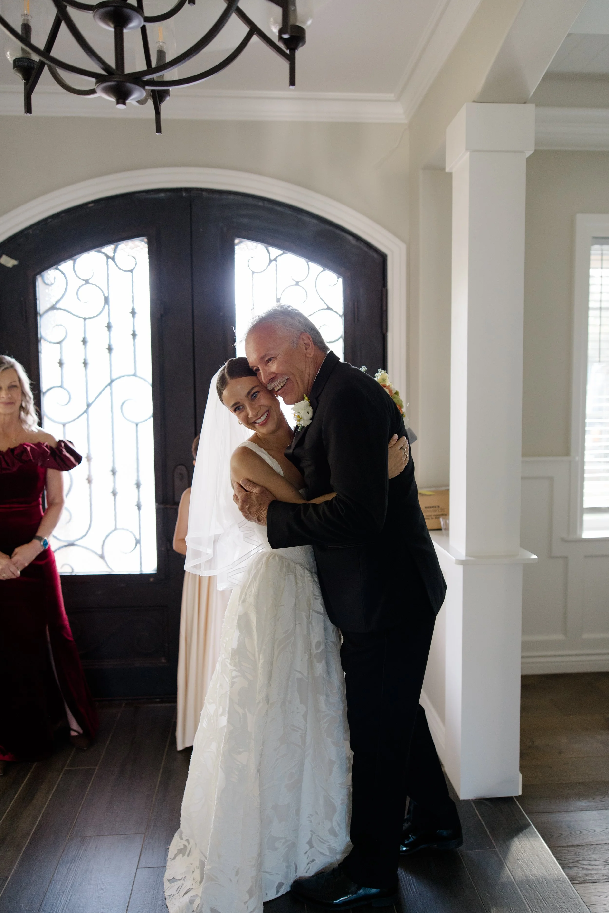 A bride and an elderly man, possibly her father, share a dance at a wedding reception inside a home, with a woman in a burgundy dress watching nearby.