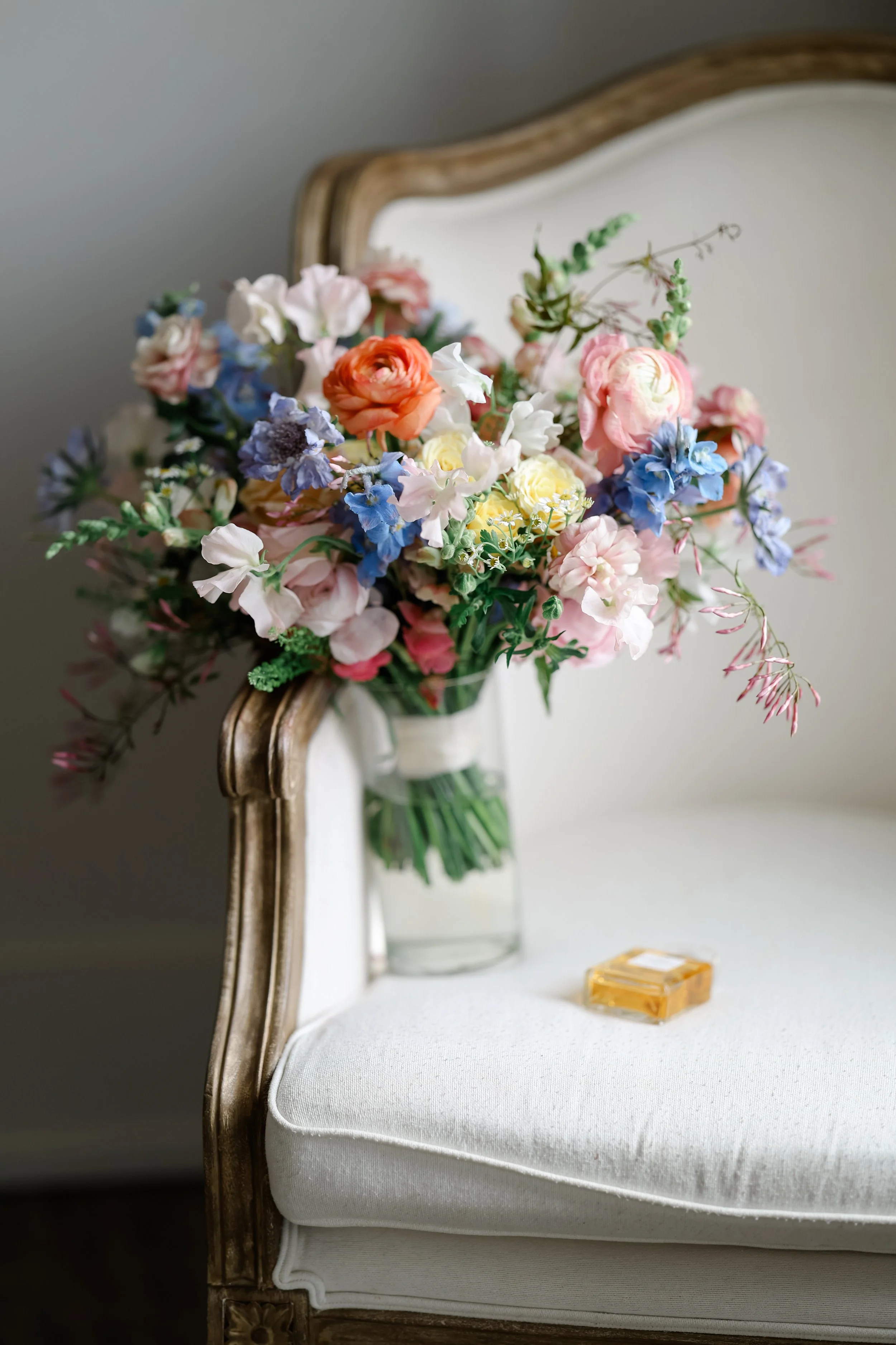 A bouquet of mixed pastel-colored flowers, including pink, purple, blue, white, and orange, sits on an antique white upholstered chair with wooden trim, next to a small perfume bottle and a gold bar of soap.