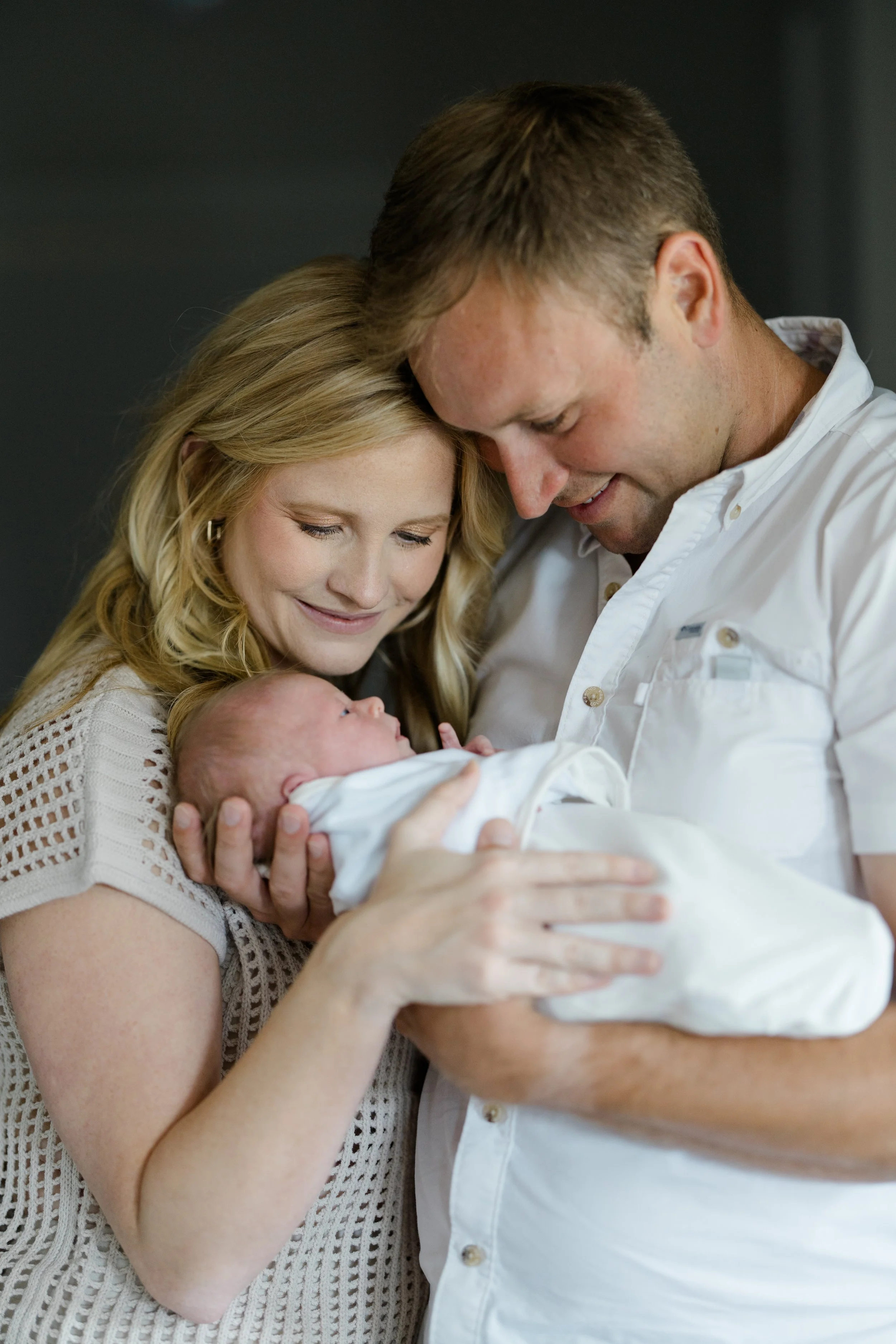 A family of three, a woman, a man, and a newborn baby, sharing a moment of affection as they look at the baby. The woman is holding the baby, and both adults are touching and leaning towards the baby with gentle smiles.