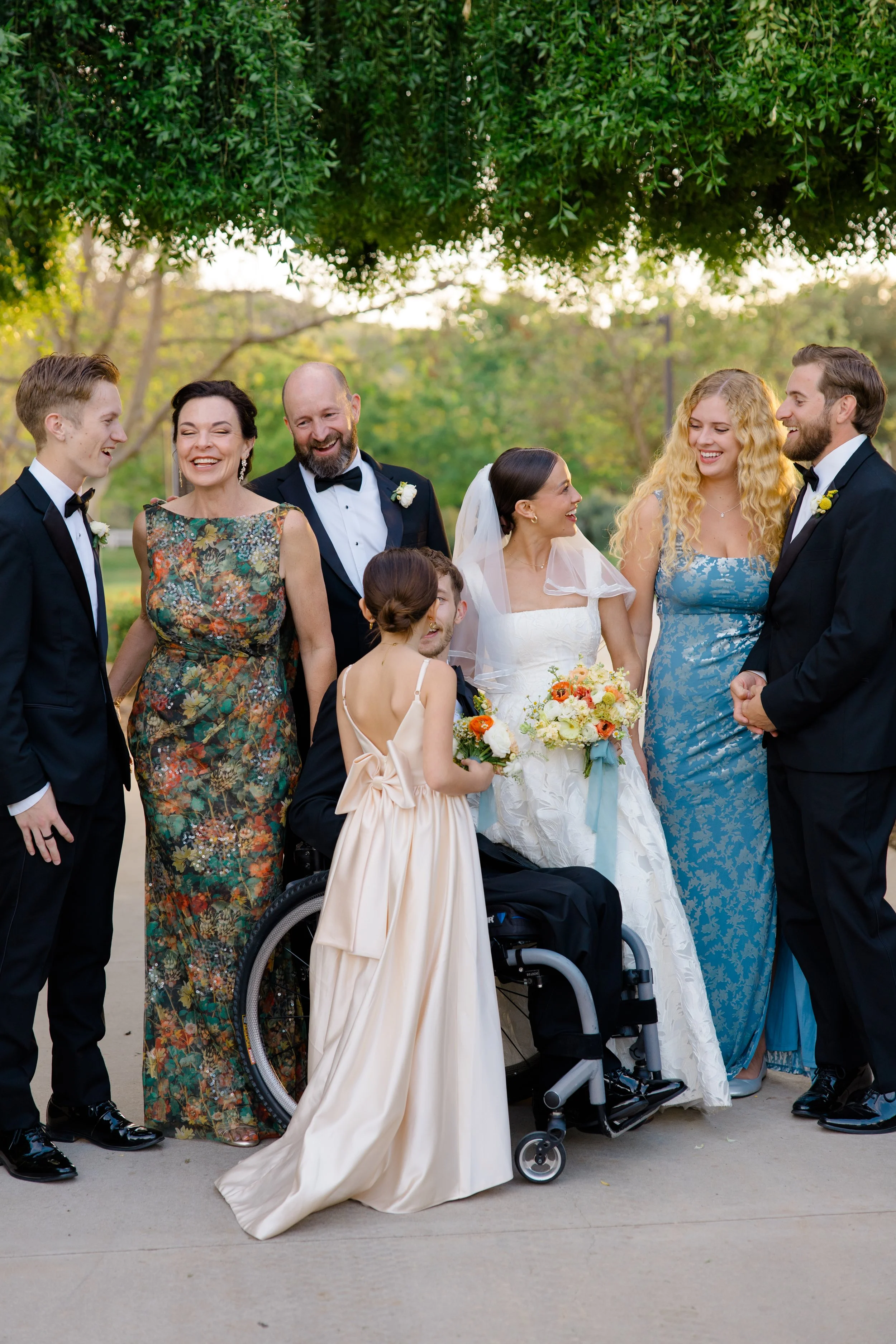 Group of people at a wedding celebration, smiling and talking outdoors, with a lush green tree overhead and a bright, natural background.