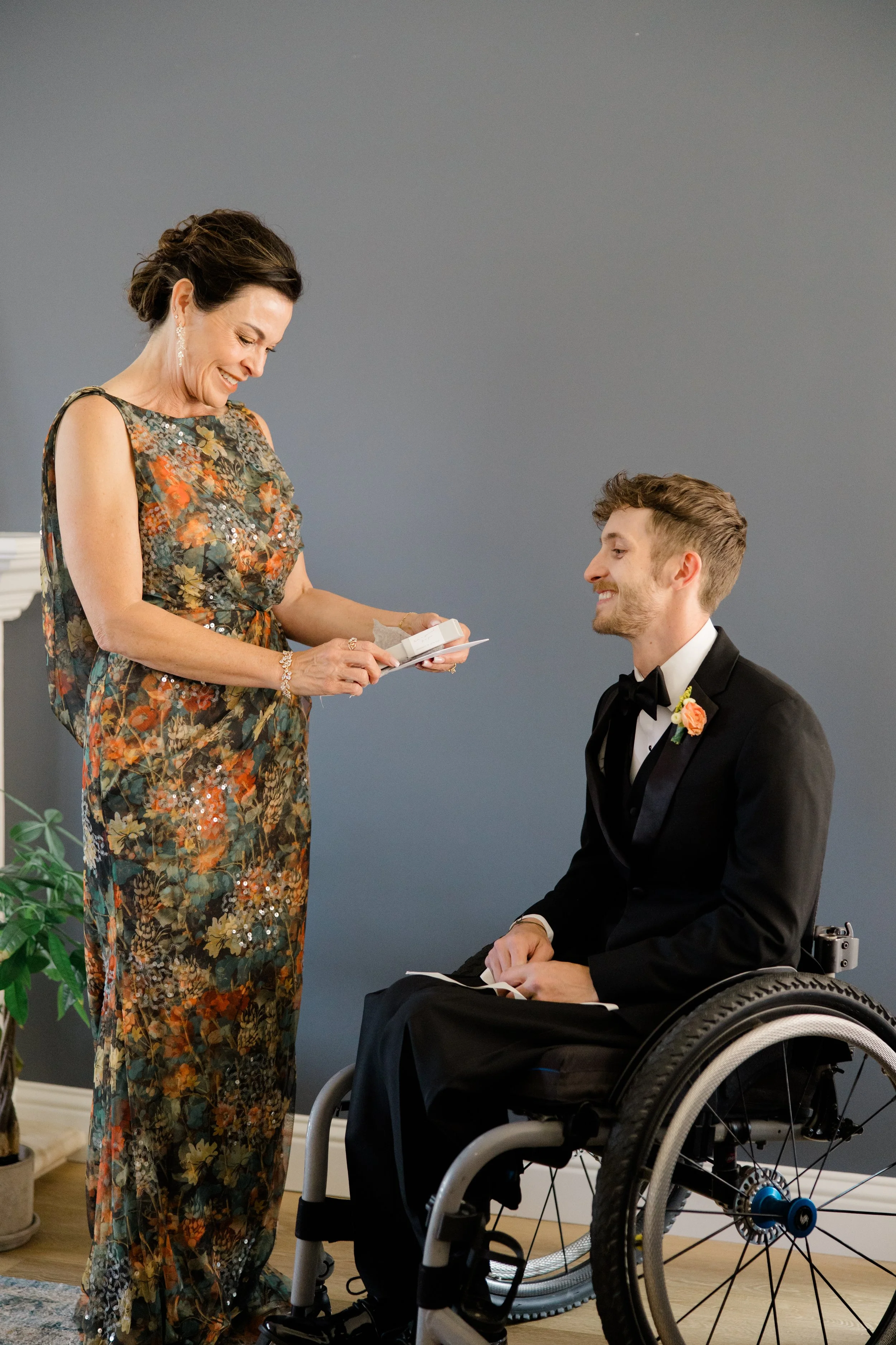 A woman in a colorful floral dress is smiling and holding a jewelry box, giving a gift to a young man dressed in a black tuxedo with a boutonniere, who is sitting in a wheelchair and smiling.