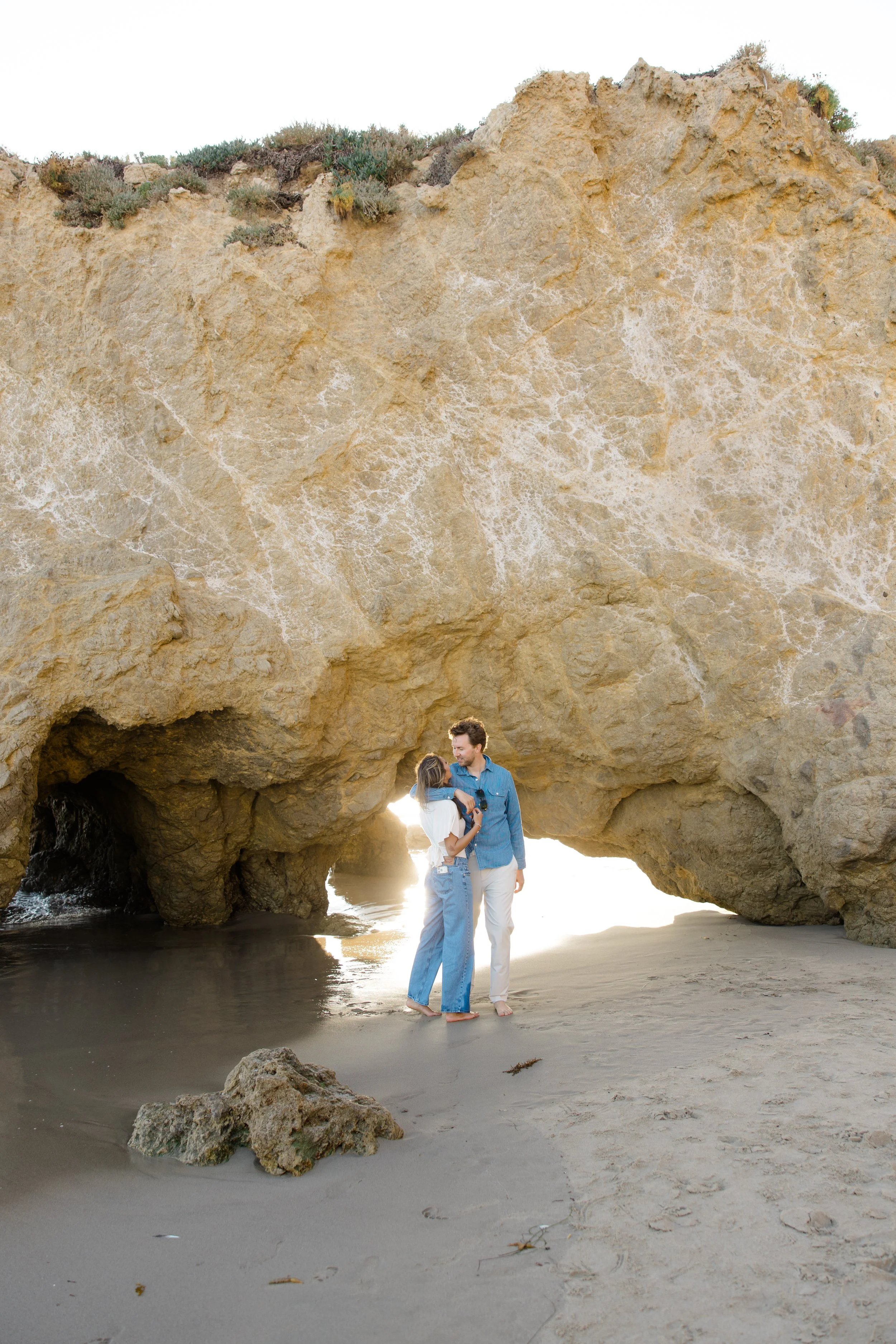 A couple standing on a sandy beach under a large natural rock arch, with sunlight shining through the opening.