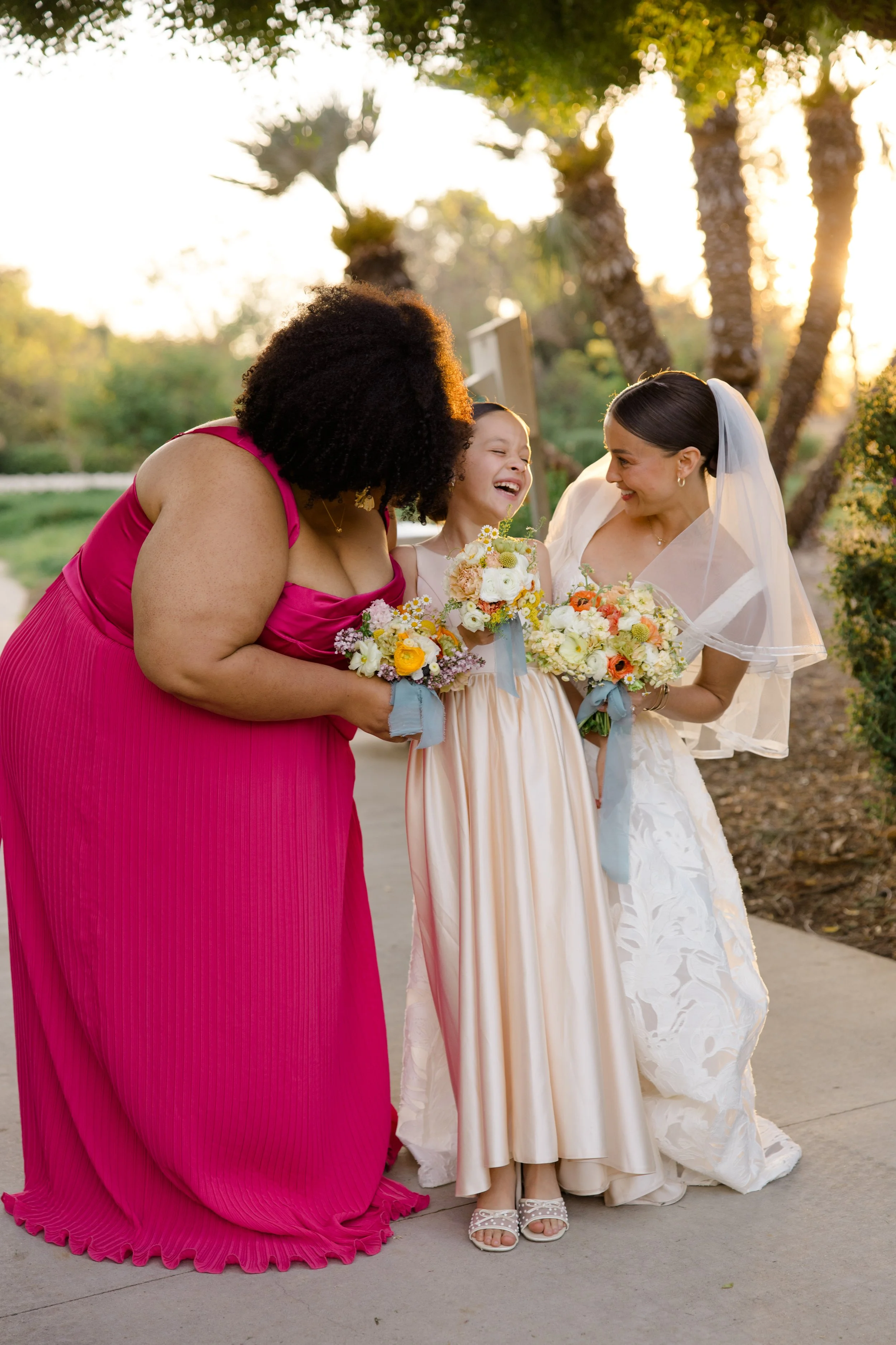 A bride in a white wedding dress, a young girl in a peach dress holding a bouquet, and a woman in a pink dress, all smiling and sharing a joyful moment outdoors at sunset surrounded by palm trees.