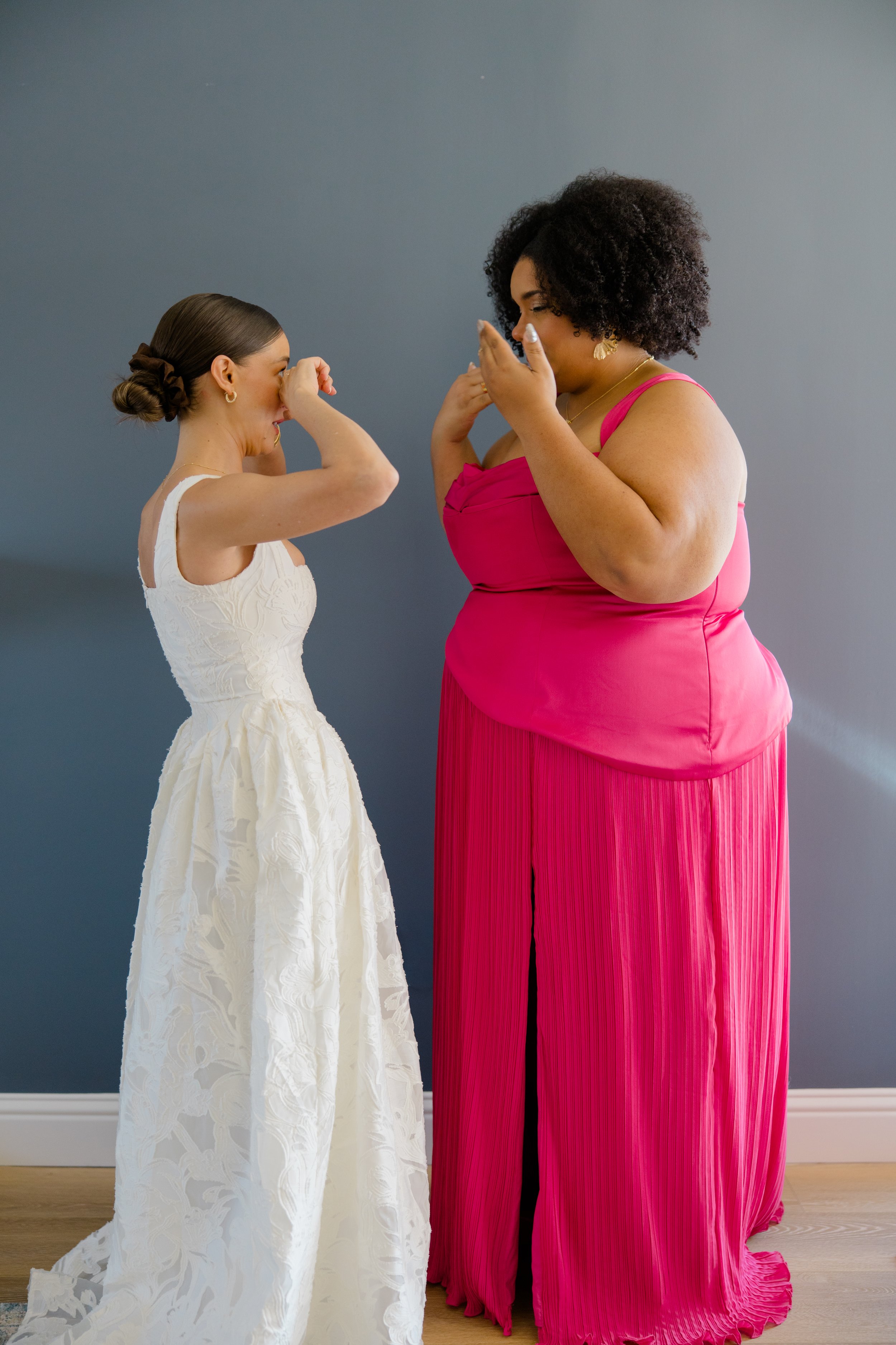 Two women are standing in front of a plain blue wall, appearing to share a joyful moment. The woman on the left is wearing a white dress with a textured floral pattern, with a bow tied at the back of her hair. She is wiping her eyes, possibly laughin