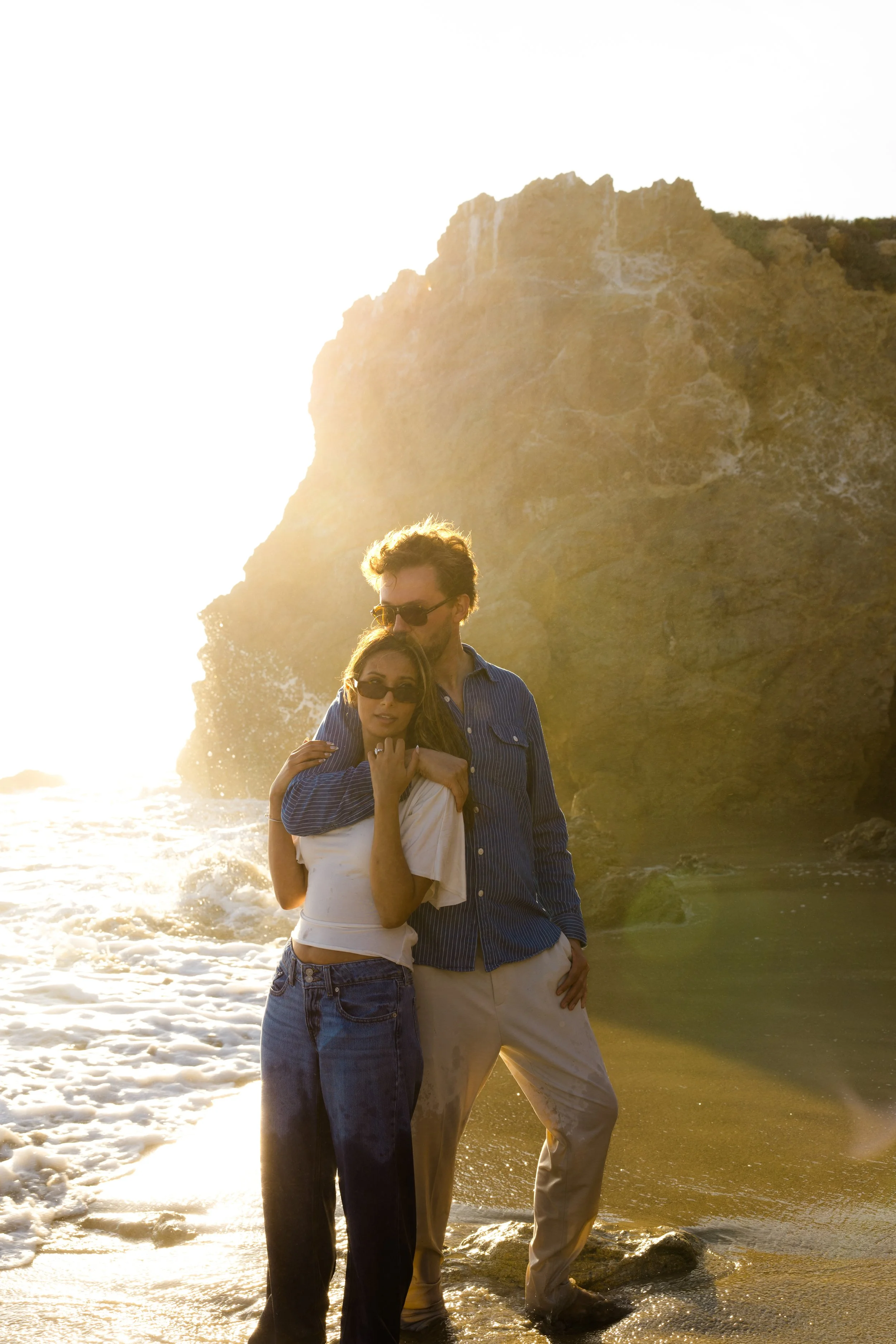 A couple standing on a beach at sunset, with large rocky cliffs in the background, one person with curly hair and sunglasses and the other with straight hair and sunglasses, embracing with the ocean waves at their feet.