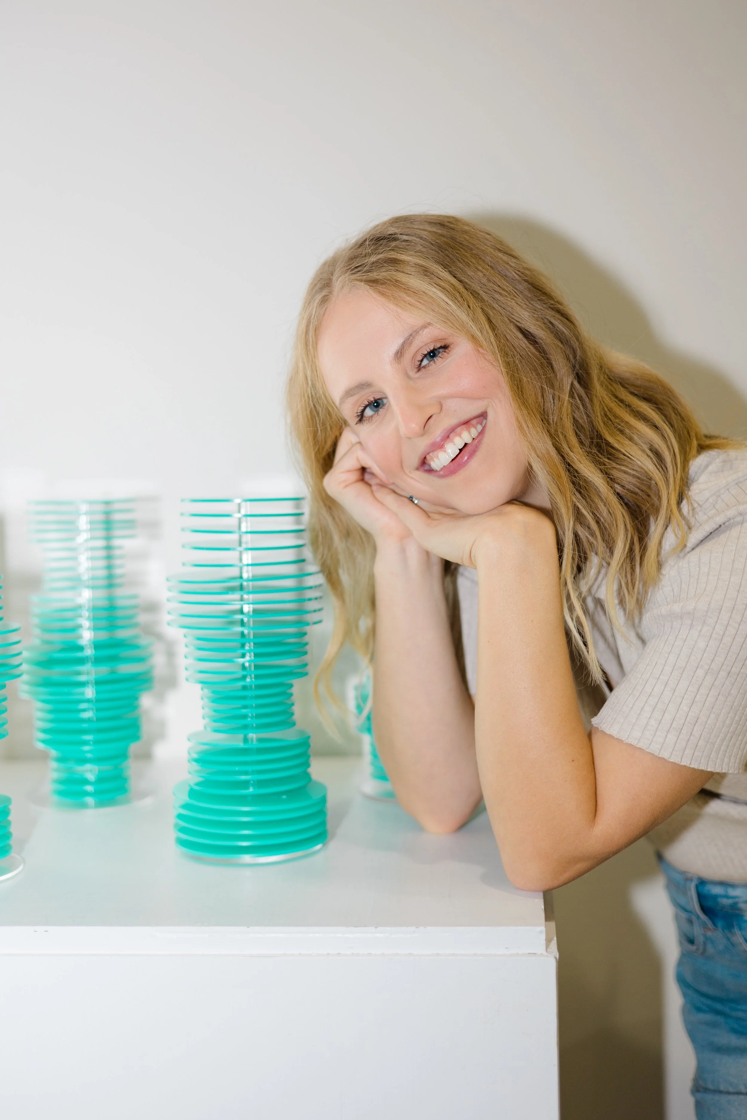 A woman with long blonde hair is smiling and resting her head on her hand while leaning on a white table. On the table, there are several vertical stacks of turquoise-colored plastic cups. The woman is wearing a light-colored, short-sleeved top and b