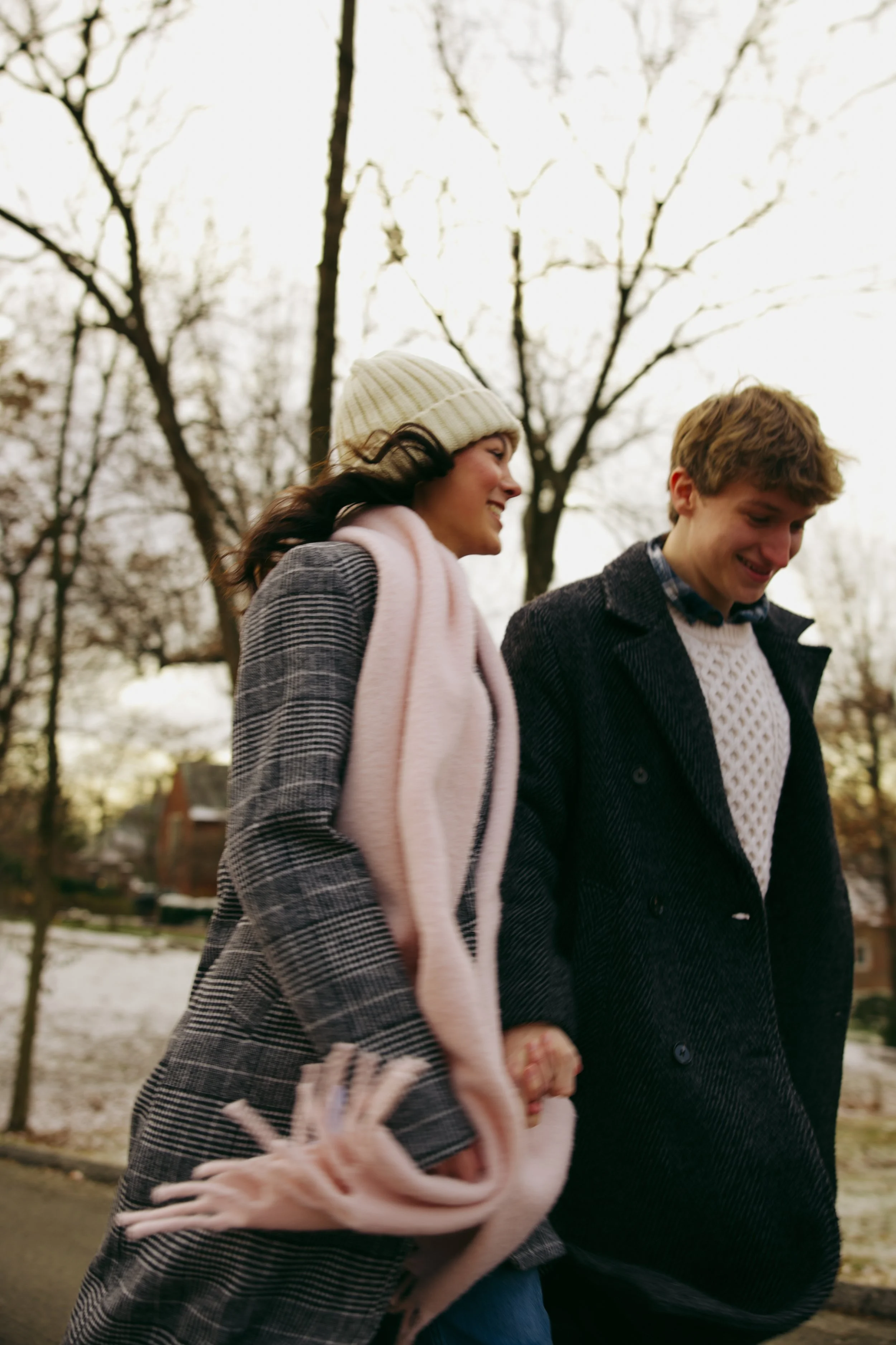 Two young people, a woman and a man, smiling and walking outdoors in fall or winter. The woman wears a cream knit hat, pink scarf, and plaid coat. The man wears a dark coat and white sweater. They hold hands, and there are leafless trees and a cloudy