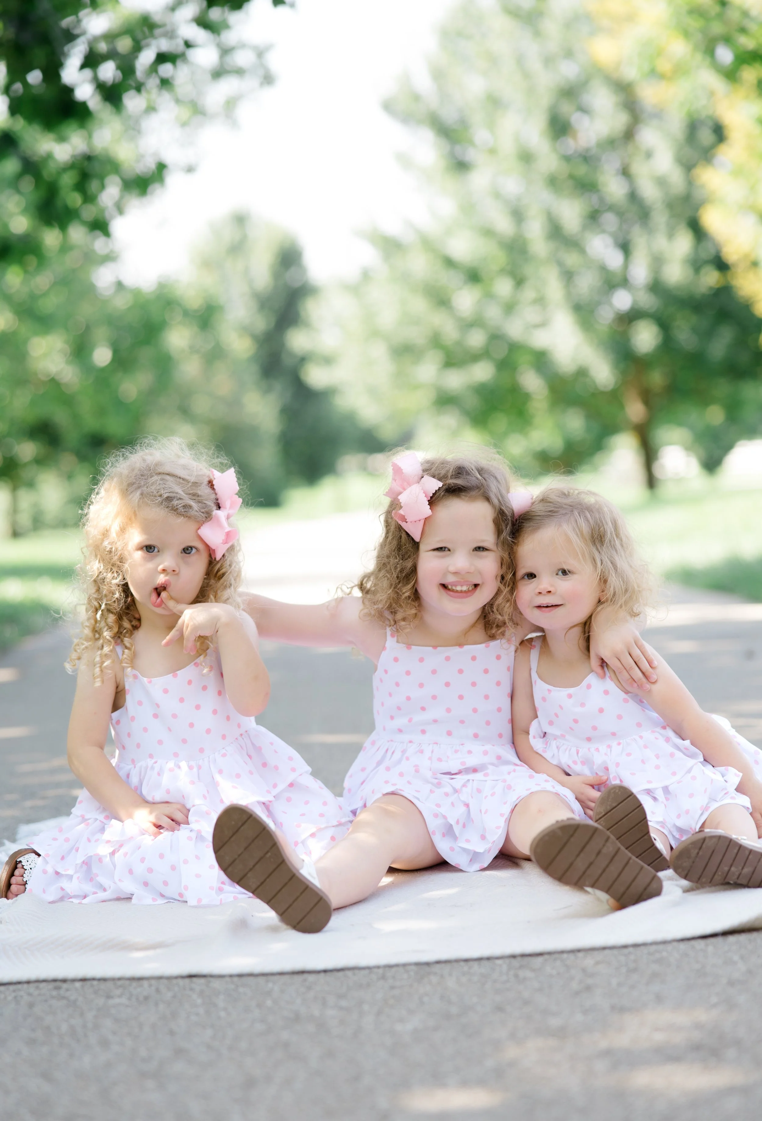 Three young girls sitting on a blanket outdoors, wearing matching pink polka dot dresses with pink bows in their hair. They are smiling and appear happy, with trees and greenery in the background.