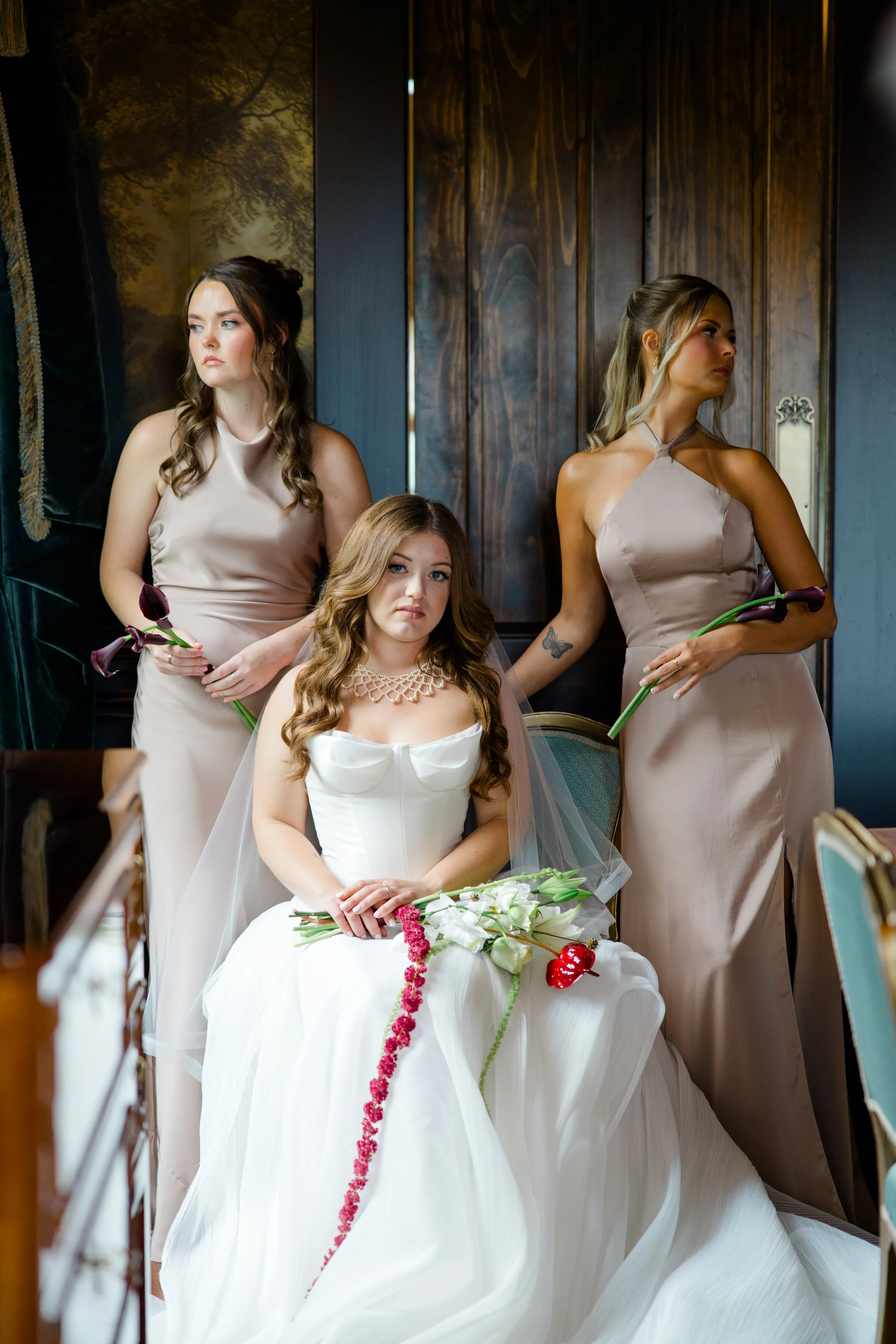 Three women in elegant dresses holding flowers sit in a formal setting, possibly a wedding or event, with dark wooden paneling and a decorative background.