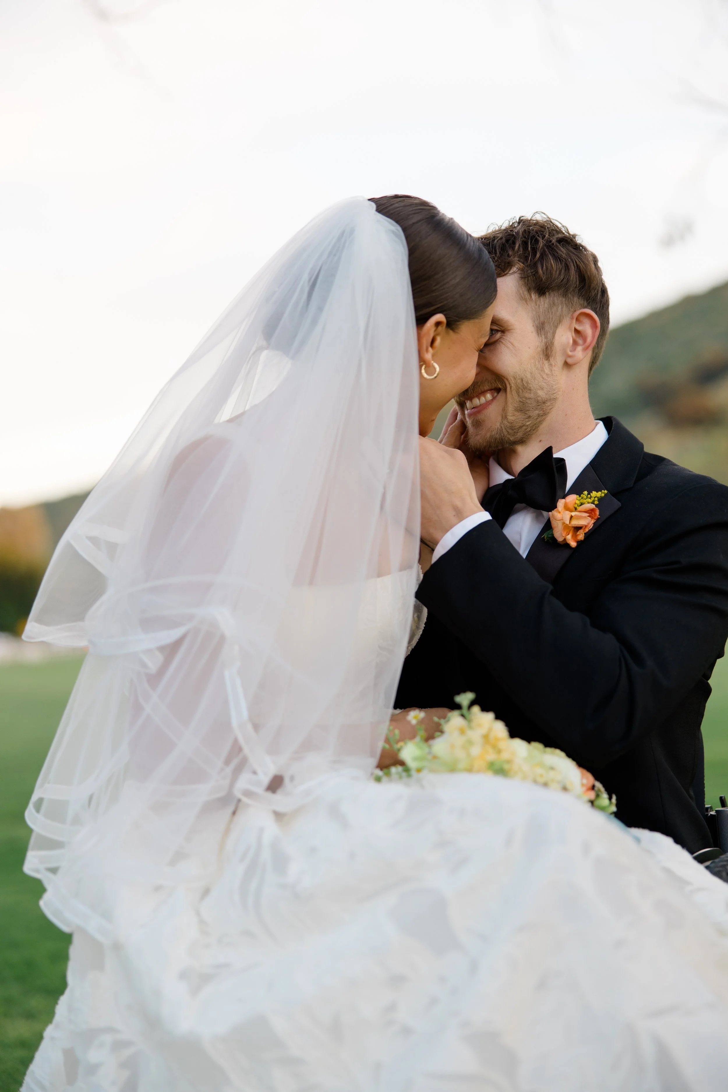 A bride and groom are smiling and touching foreheads during their wedding in an outdoor setting with hills in the background.