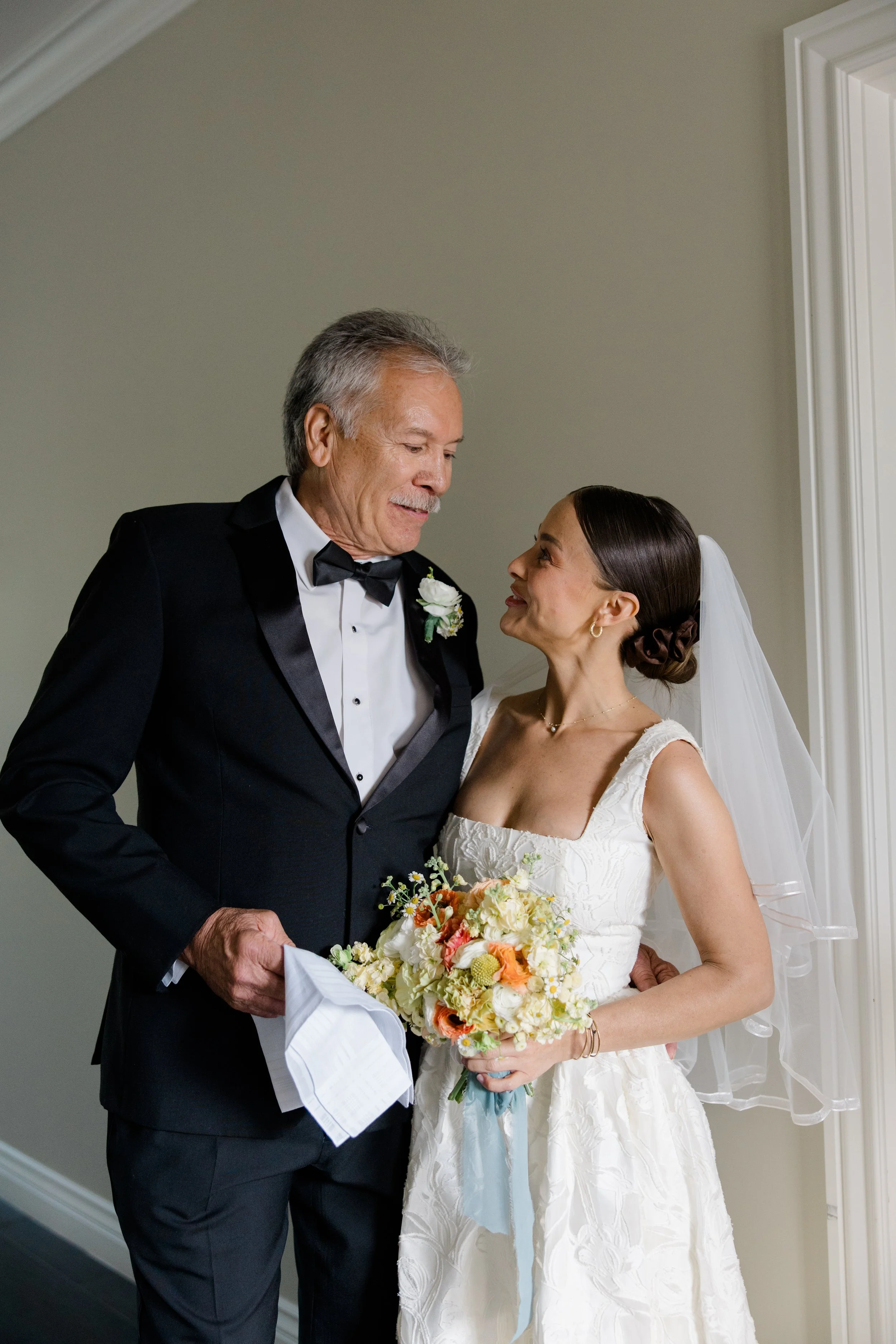 A bride and an older man, possibly her father, sharing a special moment during her wedding. The man is dressed in a black tuxedo with a bow tie, and the bride is holding a bouquet of flowers, wearing a white wedding dress and a veil. They are looking