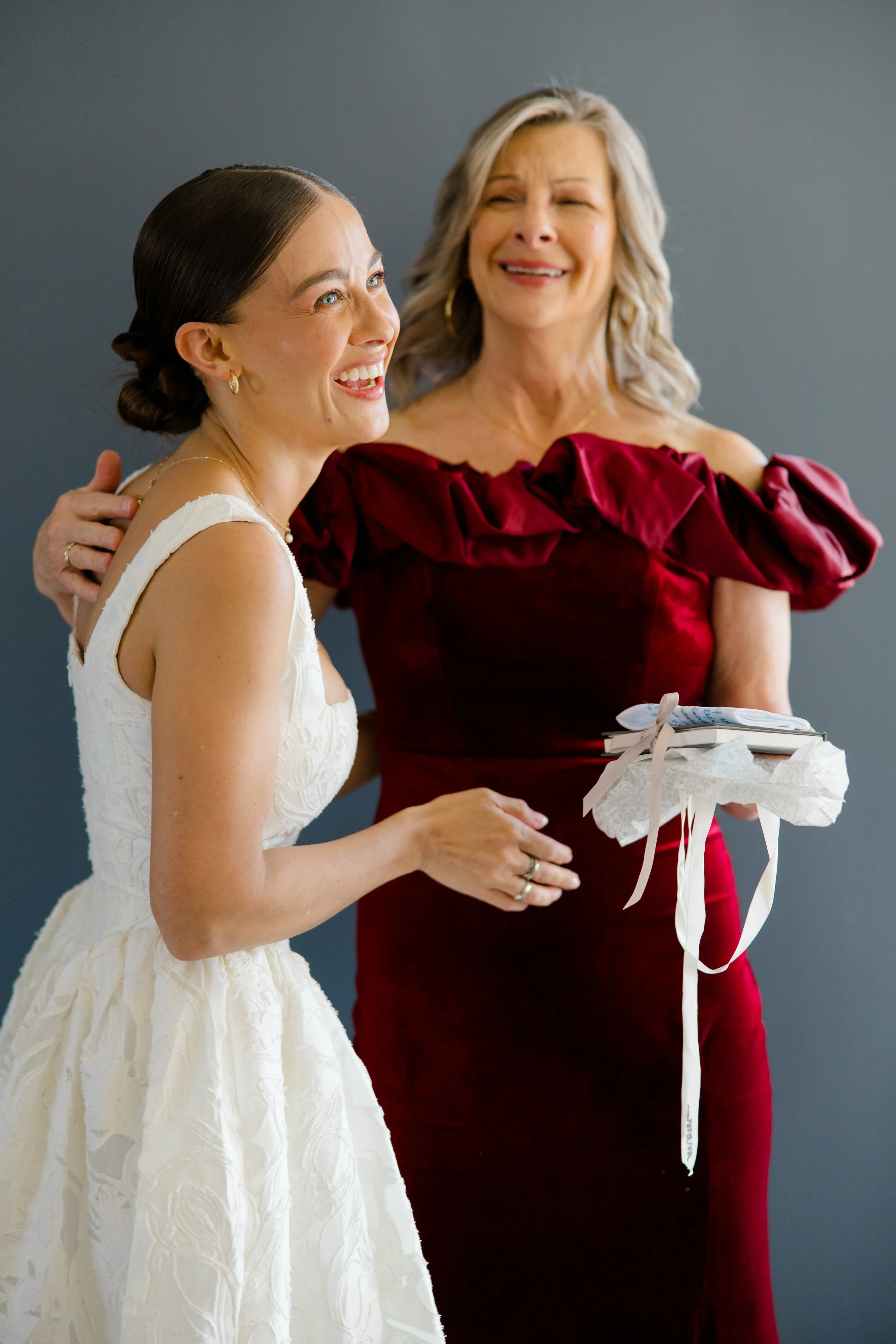 A bride smiling and looking to the side, standing next to a woman in a red dress who is holding a gift wrapped in white paper with a ribbon, against a gray background.