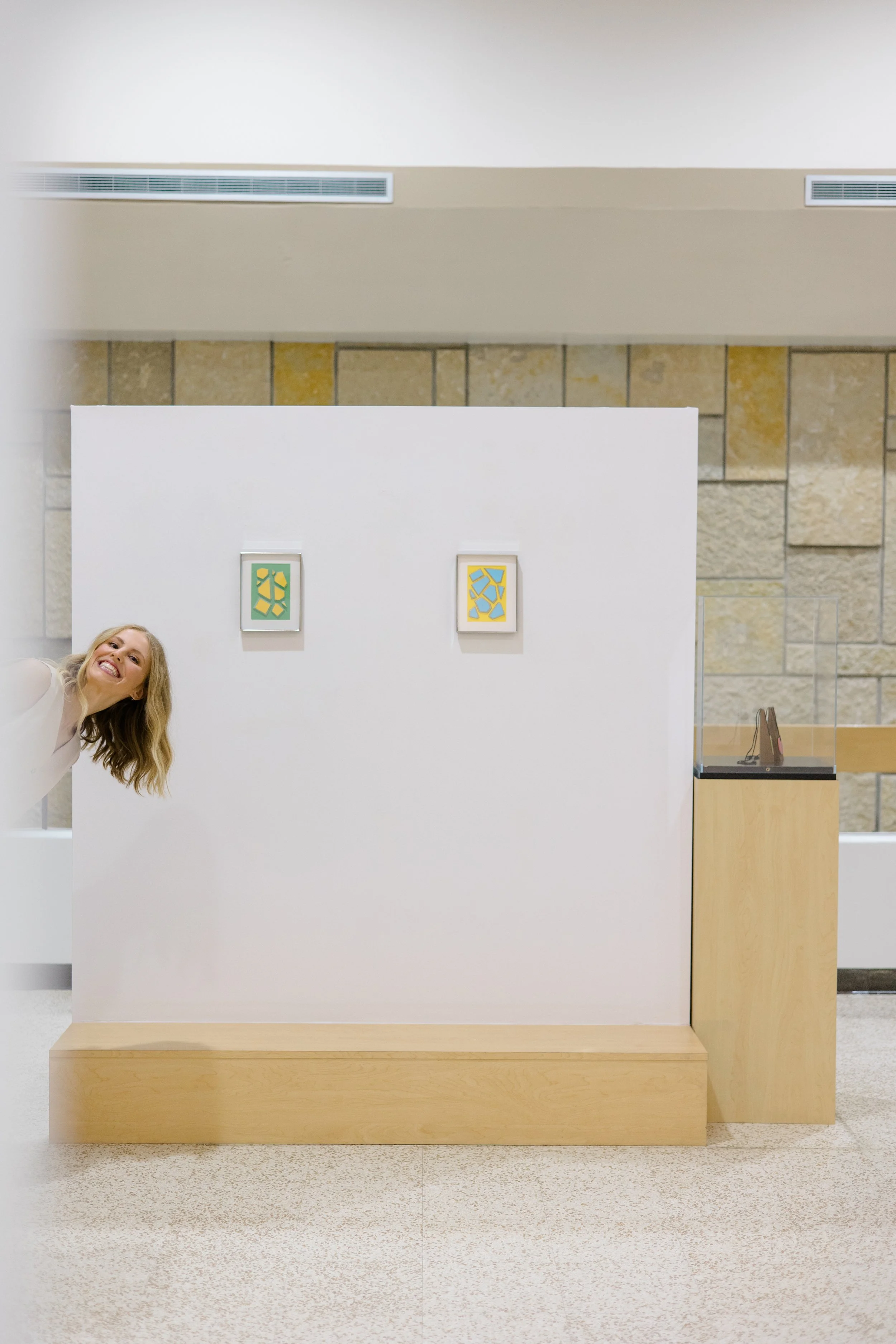 An art gallery display with two colorful abstract prints on a white wall, and a woman smiling partially visible on the left side of the image.
