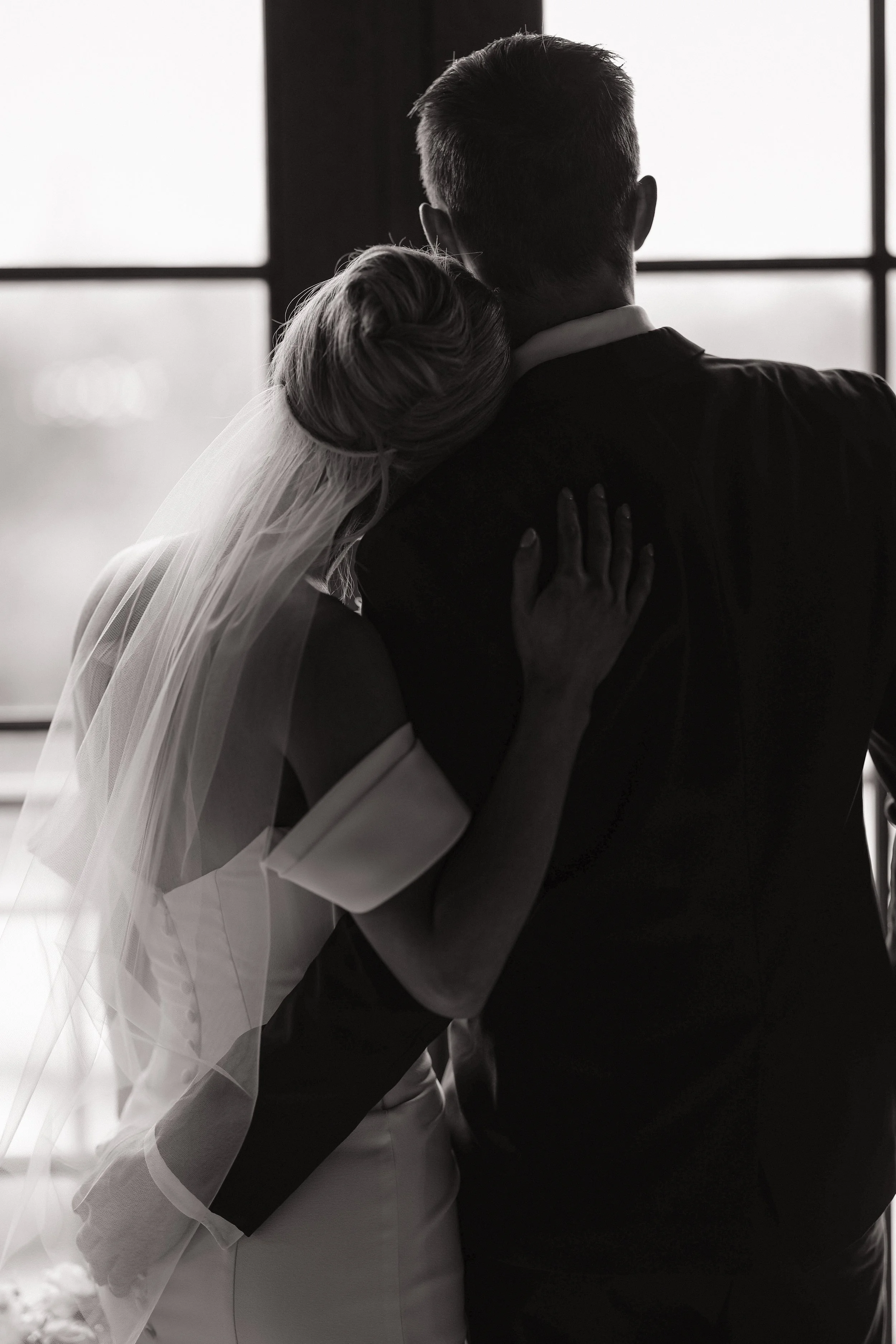 Black and white photo of a bride and groom embracing. The bride's head rests on the groom's shoulder, and her hand rests on his back. The groom's back is facing the camera, and a window is in the background.