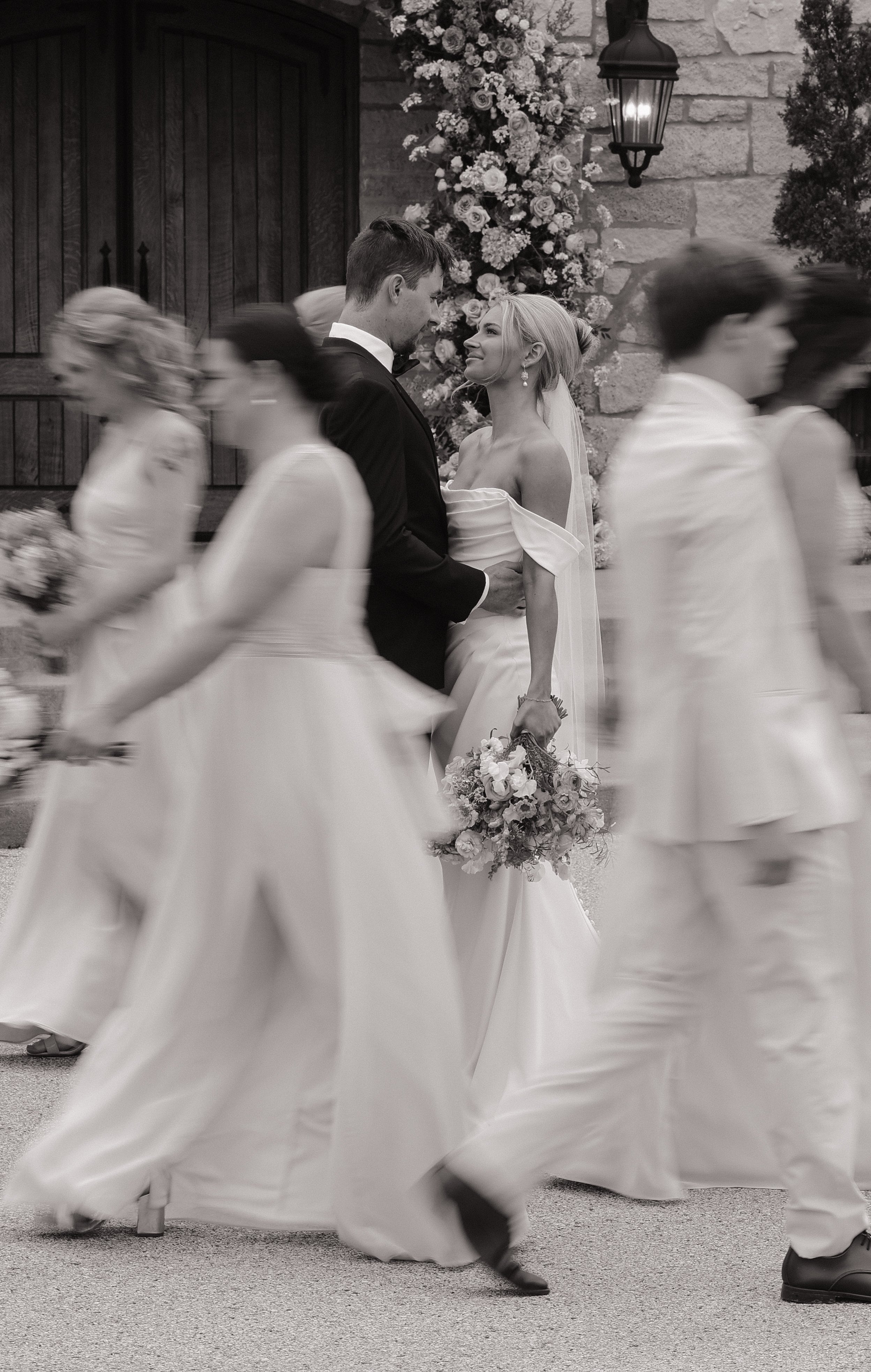 Black and white photograph of a wedding scene with a bride and groom as the central focus, surrounded by bridesmaids walking in front. The bride is holding a bouquet and is looking at the groom, who is smiling back at her, standing in front of a ston