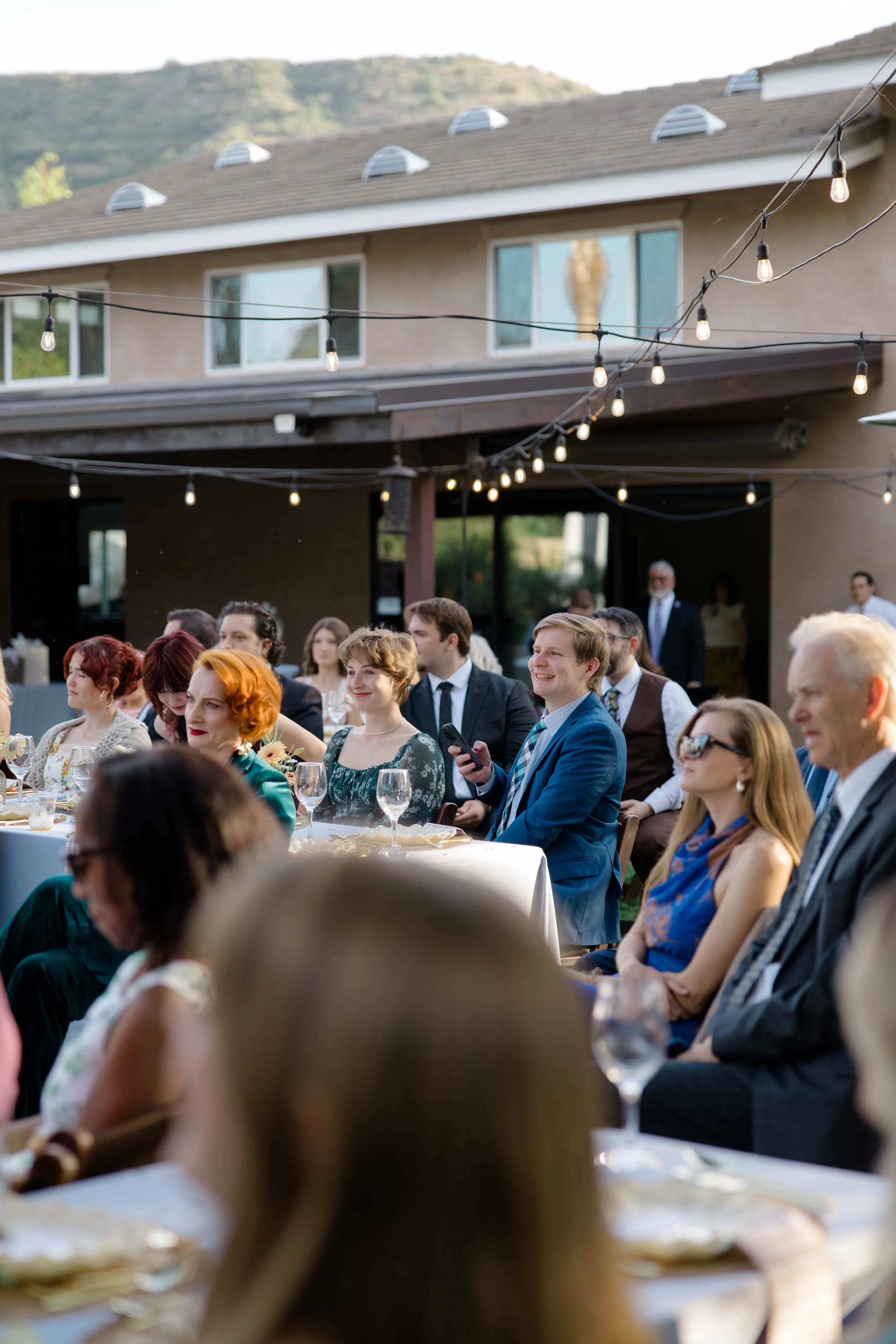 People dressed in formal attire attending an outdoor event with string lights, sitting at a long table with glasses of wine, in a backyard with a house and hills in the background.