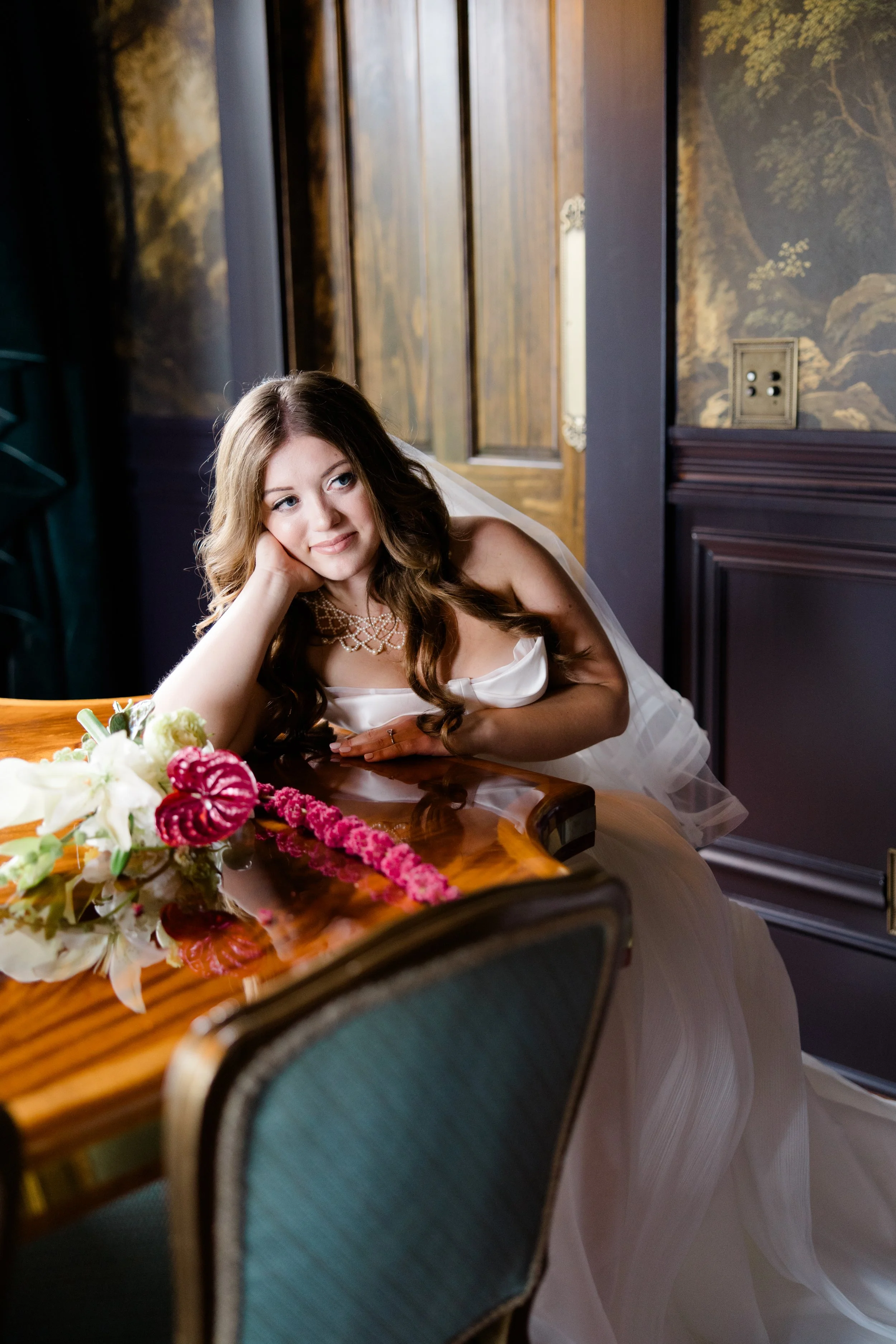 A young woman in a wedding dress with long, wavy hair resting her head on her hand, sitting at a table with floral arrangements, in an ornately decorated room with wood paneling and painted walls.