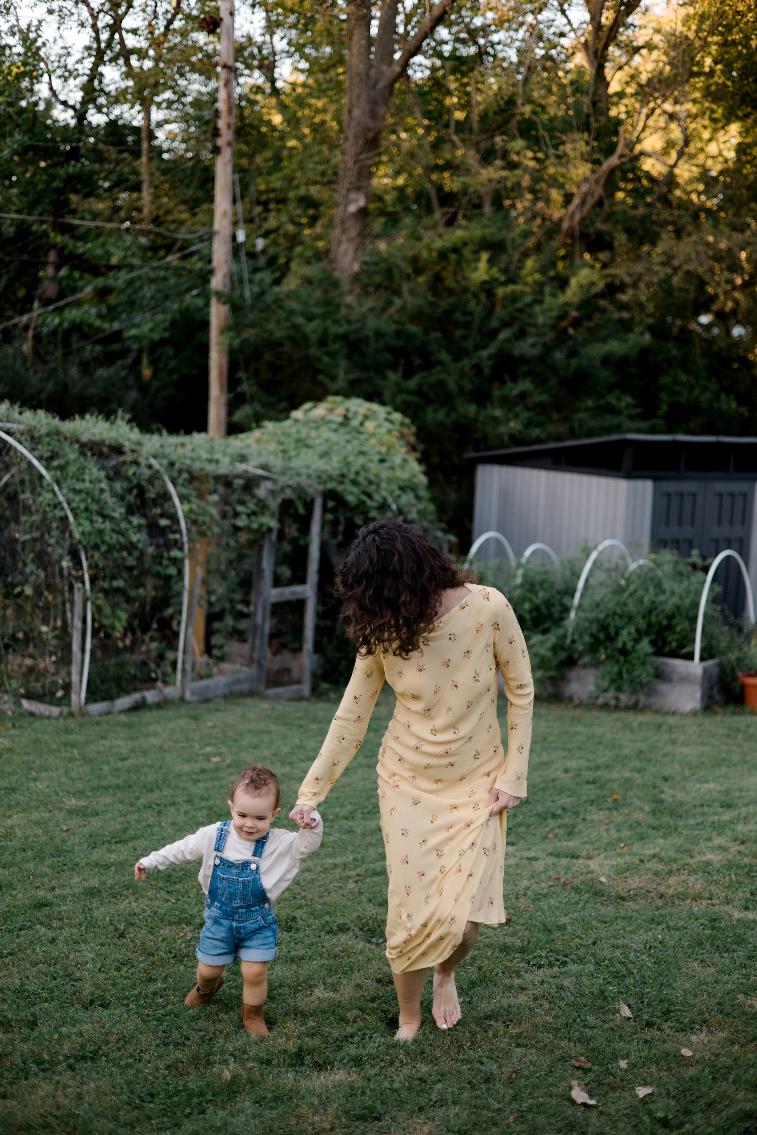 A woman in a yellow floral dress and tan sandals walking hand-in-hand with a toddler boy in denim overalls and brown boots in a backyard with green grass, trees, and a shed in the background.