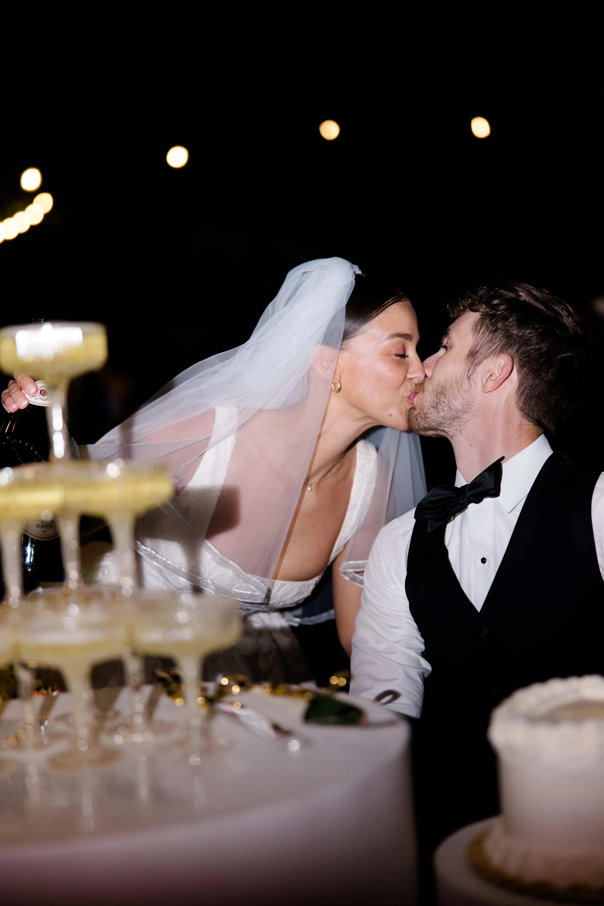 A bride and groom kissing at their wedding reception, with a wedding cake visible in the foreground.